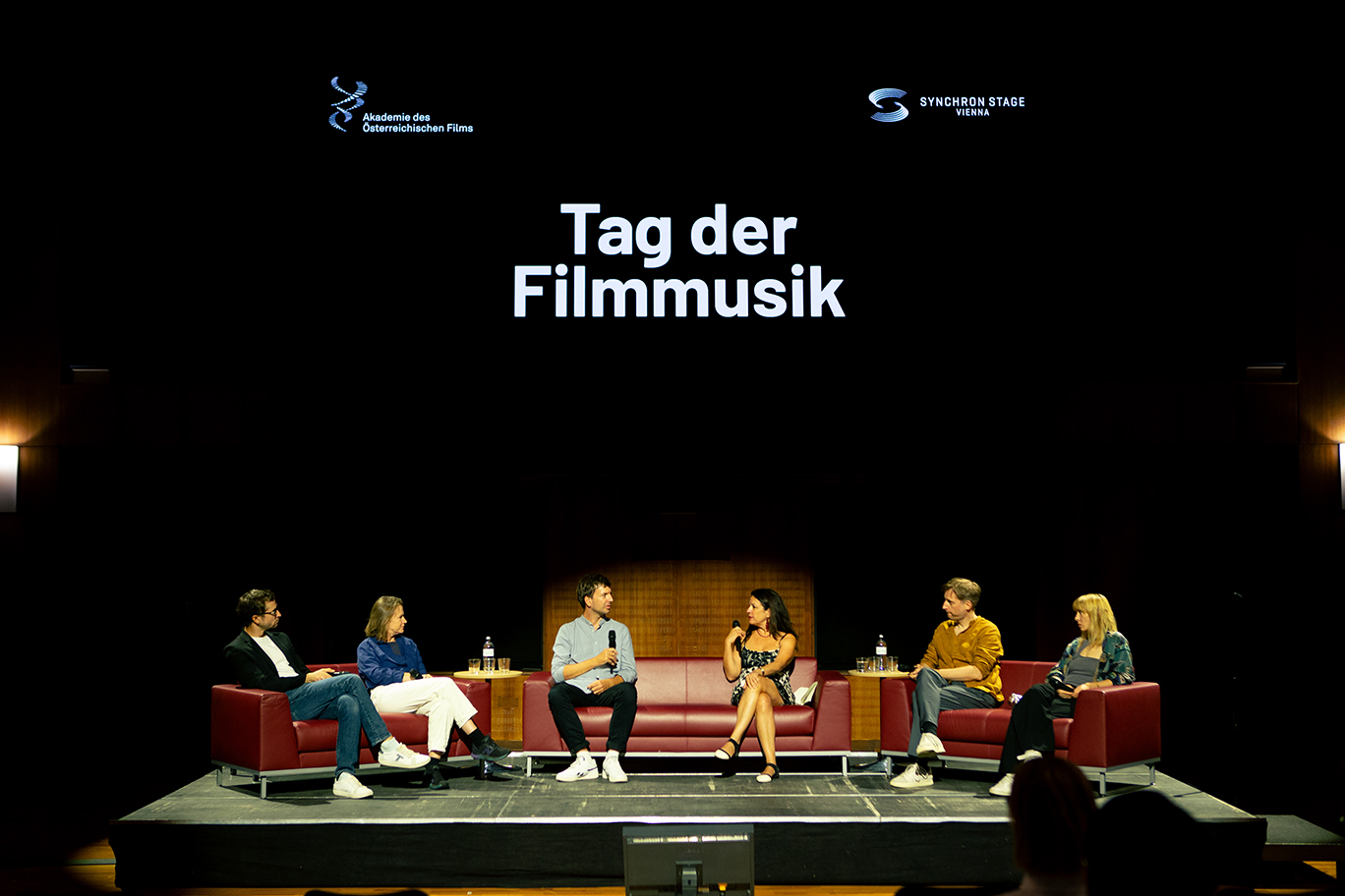 A panel of five people, including two women and three men, sit on red couches on a stage with a large screen displaying the words Tag der Filmmusik and the logos of Akademie des Österreichischen Films and Synchron Stage Vienna.