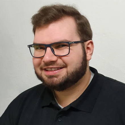 Portrait of Ben Belikov, Head of IT, a smiling man with brown hair, eyeglasses, and a beard, wearing a black polo shirt