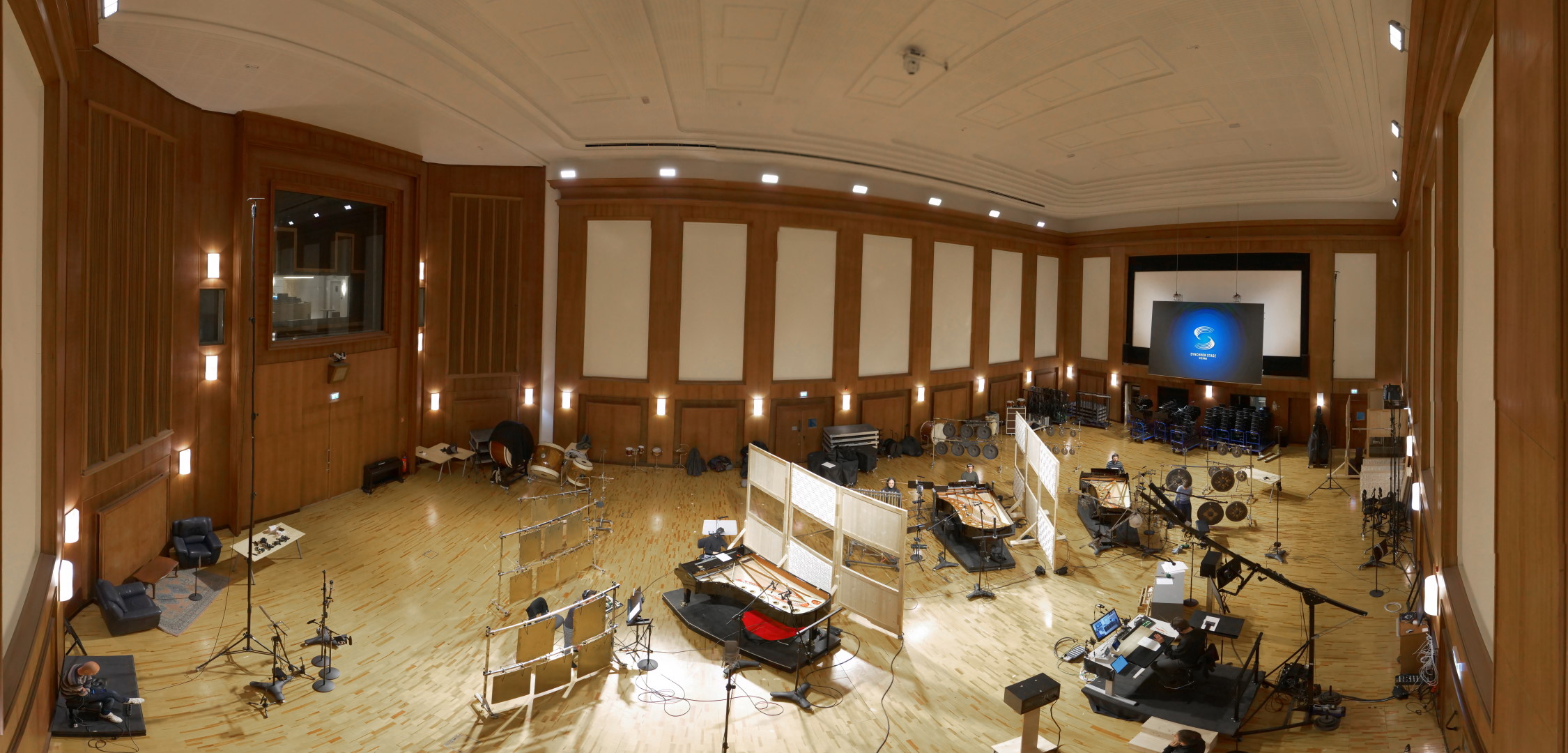 Panoramic view of the large Synchron Stage Vienna recording studio, showing orchestras setup for instruments like pianos, drums, and gongs, with sound baffling, microphones, and a control room visible through a window.