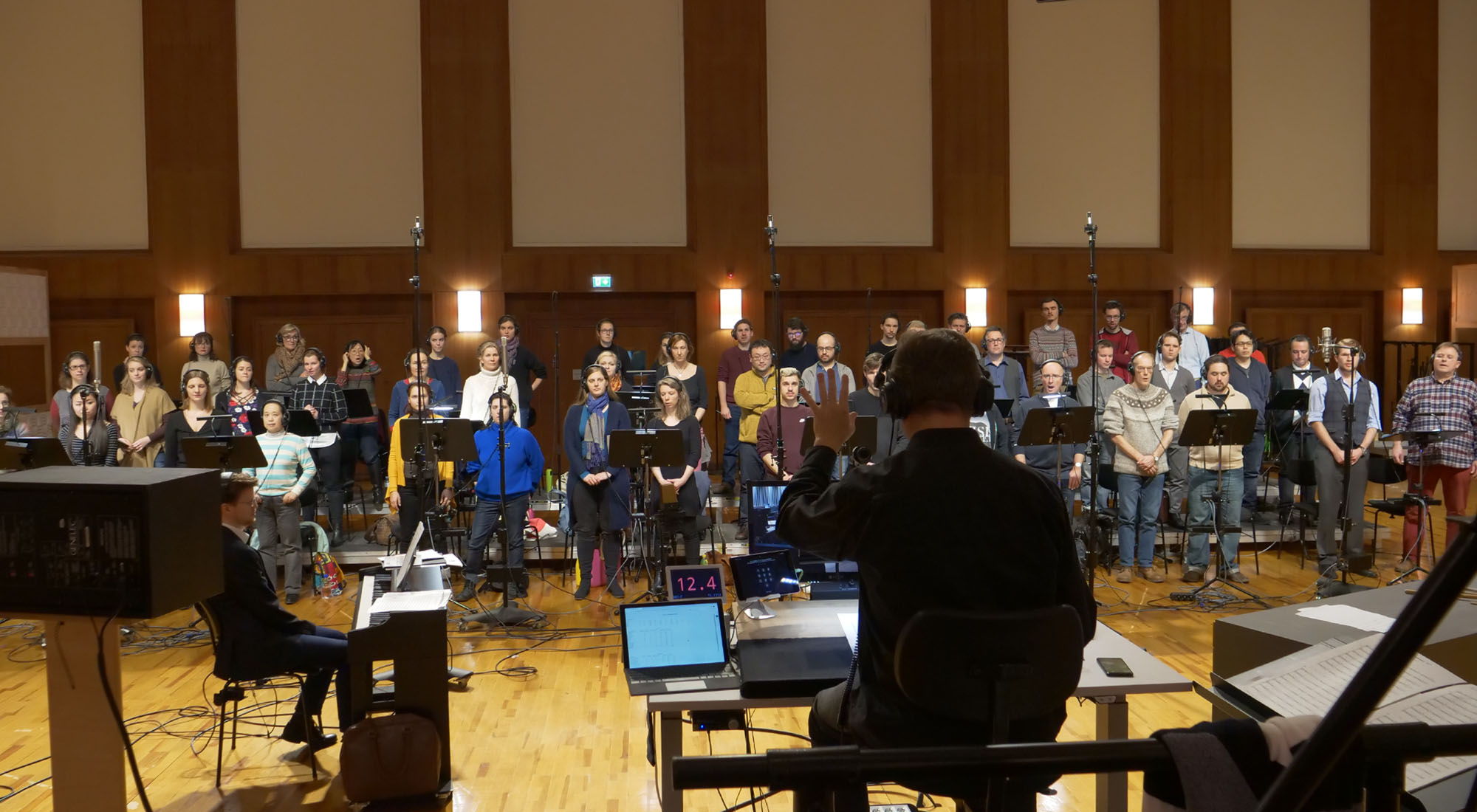 A diverse group of singers and musicians recording for the Vienna Symphonic Library's Big Bang Orchestra Ganymede album, with a conductor leading them in a professional studio setting.