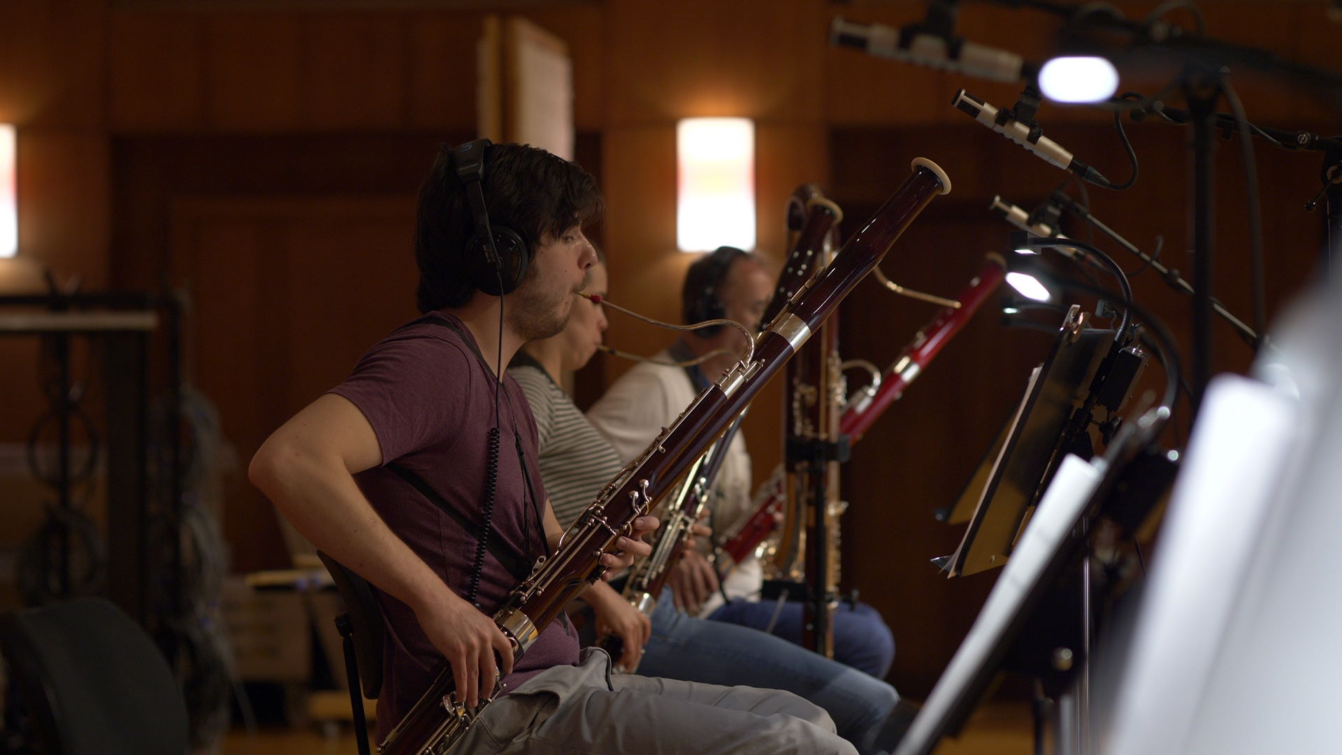 Musicians in headphones playing bassoons and other woodwind instruments in a recording studio, with microphones and music stands visible.