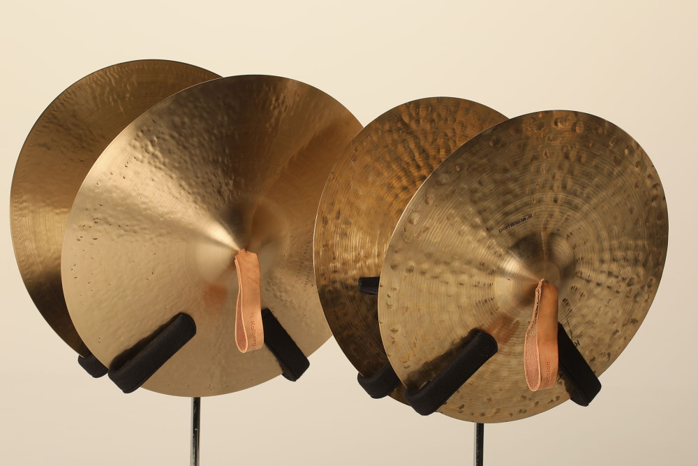 Four large orchestral cymbals, two pairs, each with a tanned leather strap for holding, displaying their hammered bronze texture.
