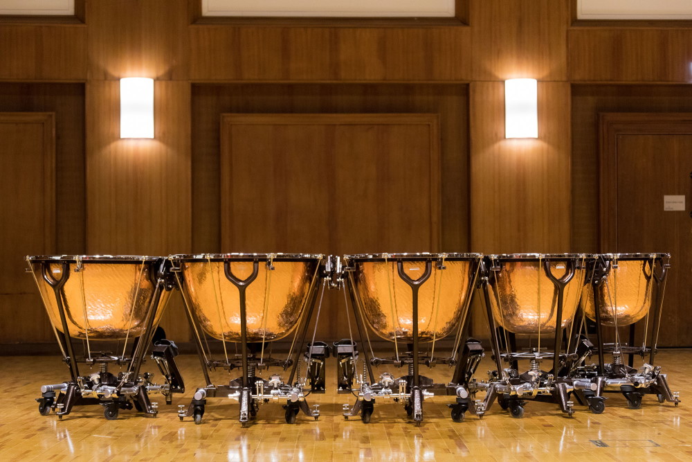 Four copper timpani, also known as Pauken or kettle-drums, with light-colored heads, on dark stands, arranged in a row against a black background.