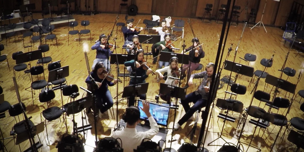 Dynamic overhead shot of the Synchron Strings I orchestra recording violins and cellos in a large studio, with microphones and music stands set up on a light wooden floor for Vienna Symphonic Library instruments.