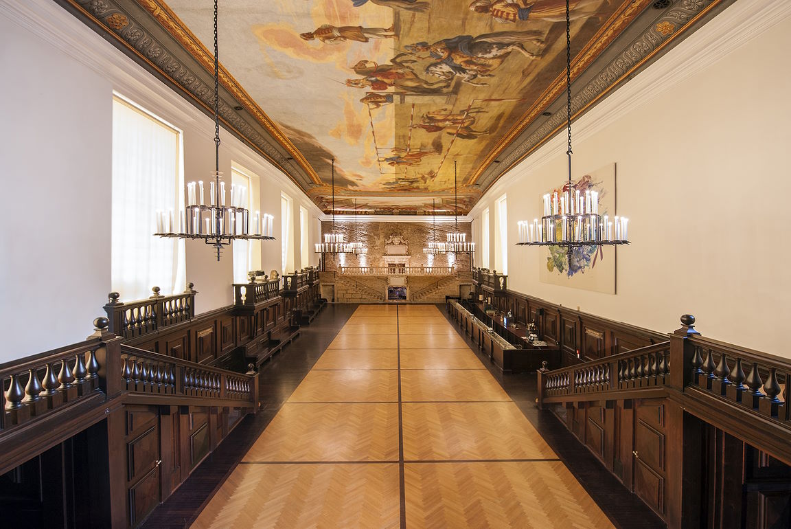 Grand hall foyer of the Grosses Festspielhaus Salzburg, featuring a long parquet floor, ornate wooden banisters, two large chandeliers, and a detailed painted ceiling.