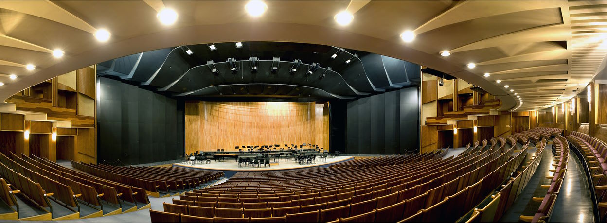 Panoramic view of the Grosses Festspielhaus concert hall in Salzburg, featuring rows of empty wooden and upholstered seats, a large stage with musical instruments and chairs, and a curved, dark ceiling with recessed lights.