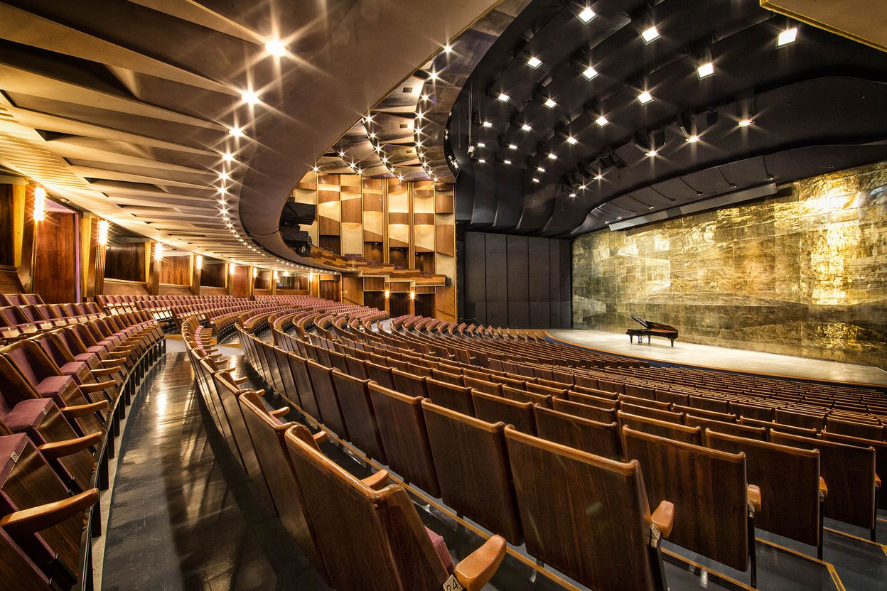 Interior view of the Grosses Festspielhaus Salzburg, showcasing rows of empty brown theater seats, a grand piano on a large stage with a detailed golden textured wall, and unique ceiling lights and architectural accents.