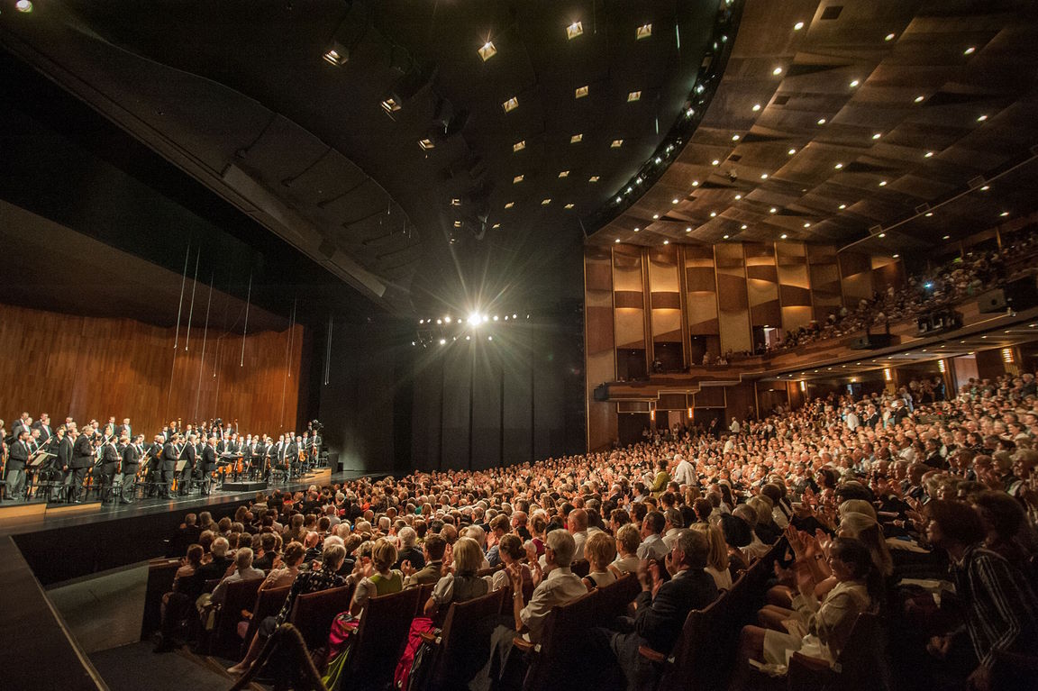 Wide shot of the Grosses Festspielhaus Salzburg interior, showing a large audience filling an auditorium facing an orchestra performing on stage under bright spotlights.