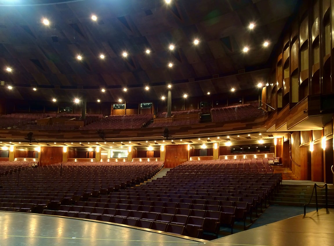 Interior view of the Grosses Festspielhaus Salzburg, an empty concert hall with rows of purple seats, wooden acoustic paneling, and lit ceiling spotlights