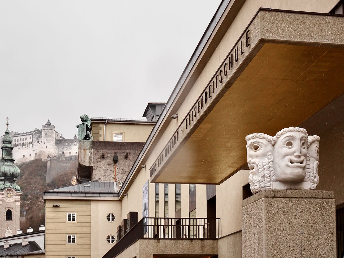 Grosses Festspielhaus Salzburg exterior, showing the modern building with ancient Greek theater mask sculpture, Hohensalzburg Fortress on a hill, and a church steeple under a cloudy sky.