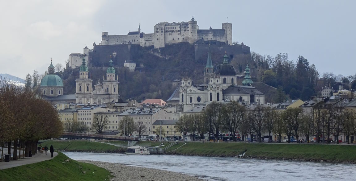 Panoramic view of Salzburg, Austria, featuring the historic Hohensalzburg Fortress on a hill overlooking the Salzach River, the city, and the Grosses Festspielhaus.