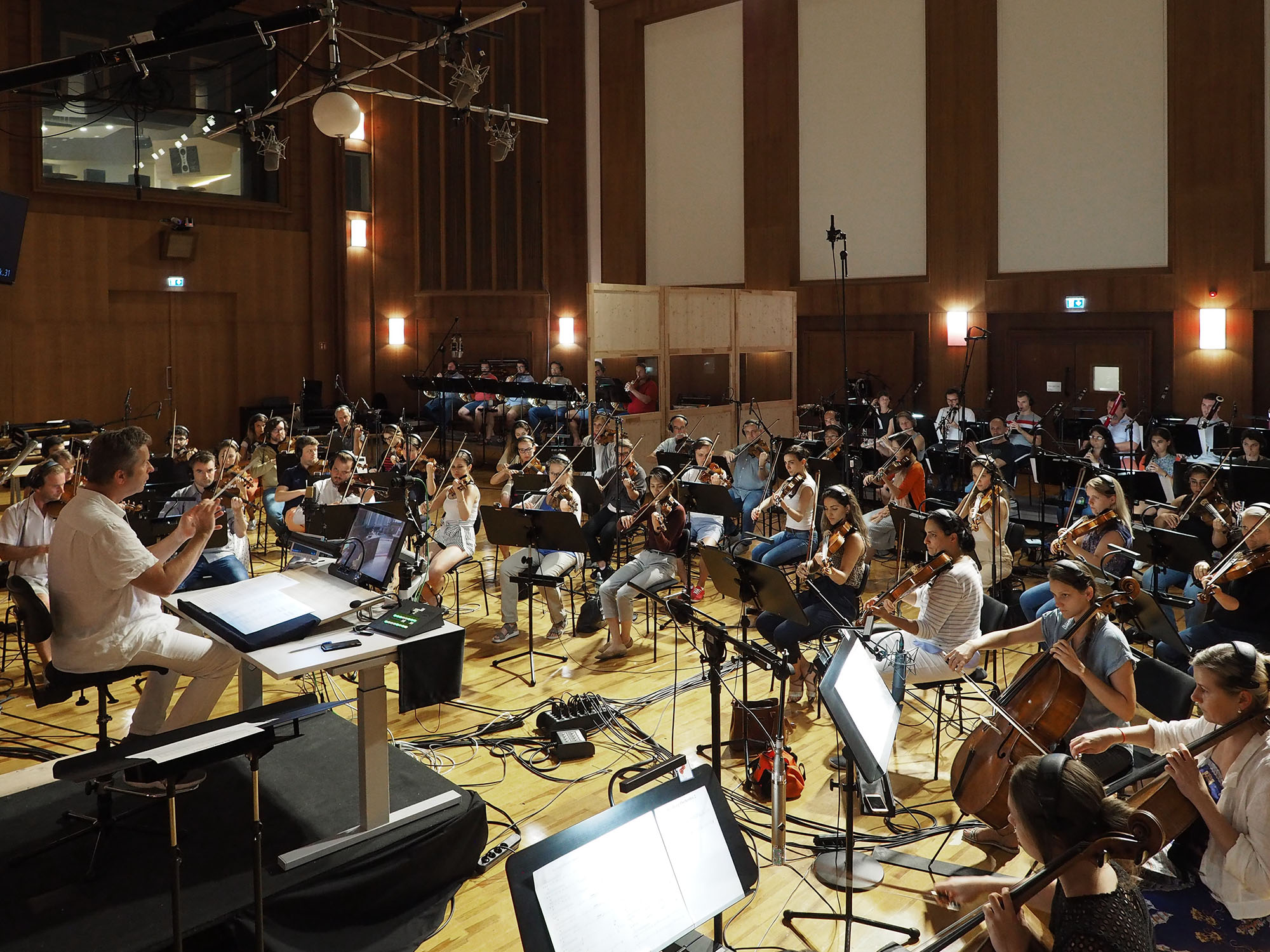 Full orchestra performing in a professional recording studio with wooden walls and acoustic panels, featuring string, woodwind, and brass sections, a conductor on a riser, and various microphones and stands setup for audio recording.