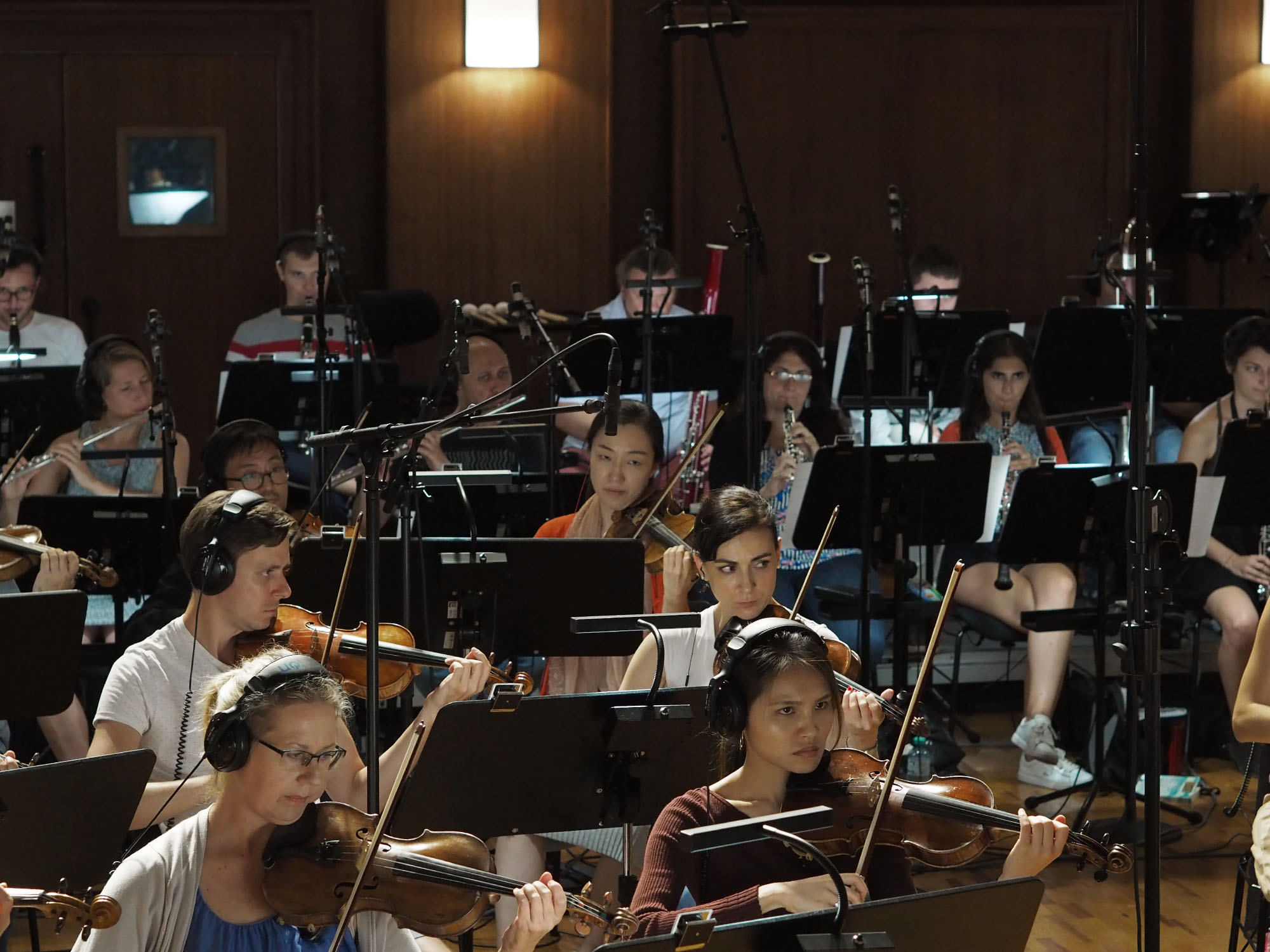 Diverse group of musicians performing in a recording studio, featuring violinists, flutists, and bassoonists, some wearing headphones, with music stands and microphones visible.