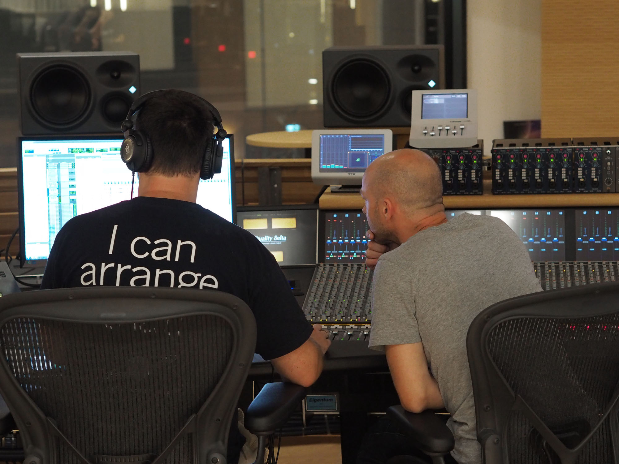 Two audio engineers in a sound mixing studio, one in headphones works at a computer monitor and audio equipment, the other watches.