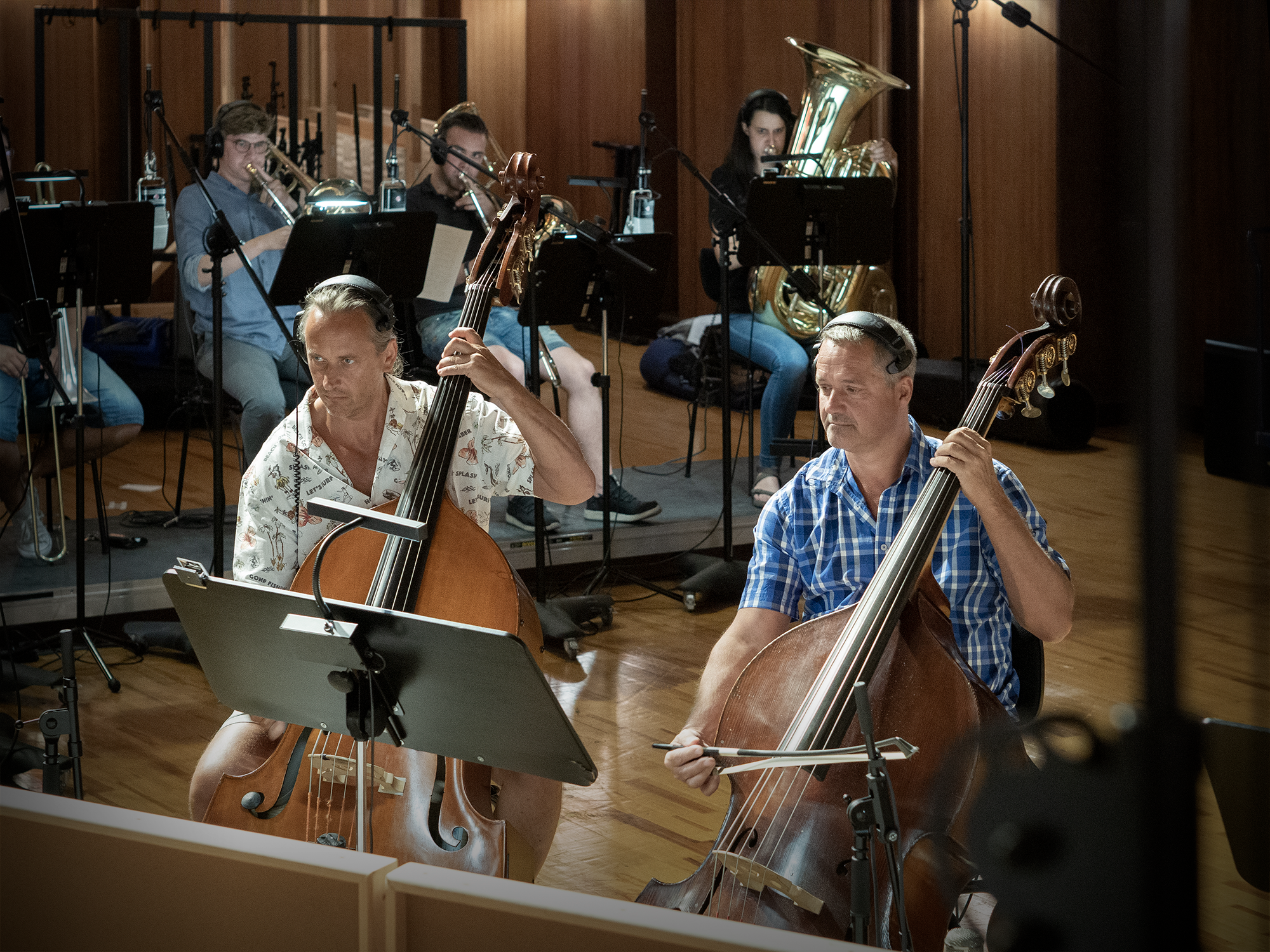 Musicians perform an orchestra in a recording studio, with two men playing upright basses in the foreground and other musicians with a trombone and tuba in the background.
