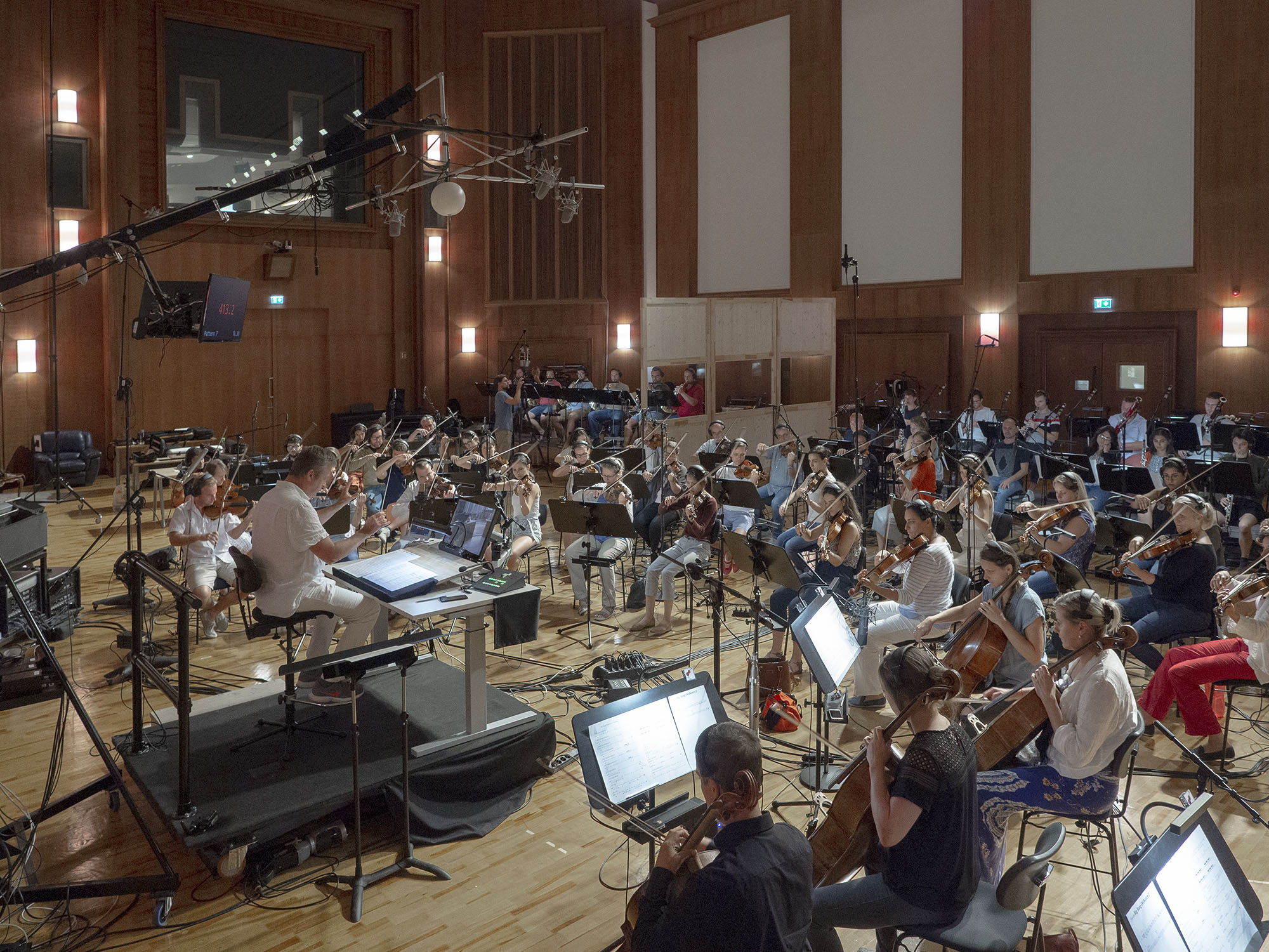 Full orchestra performing in a recording studio, featuring violinists, cellists, and musicians playing various instruments surrounded by audio recording equipment, microphones, and music stands.