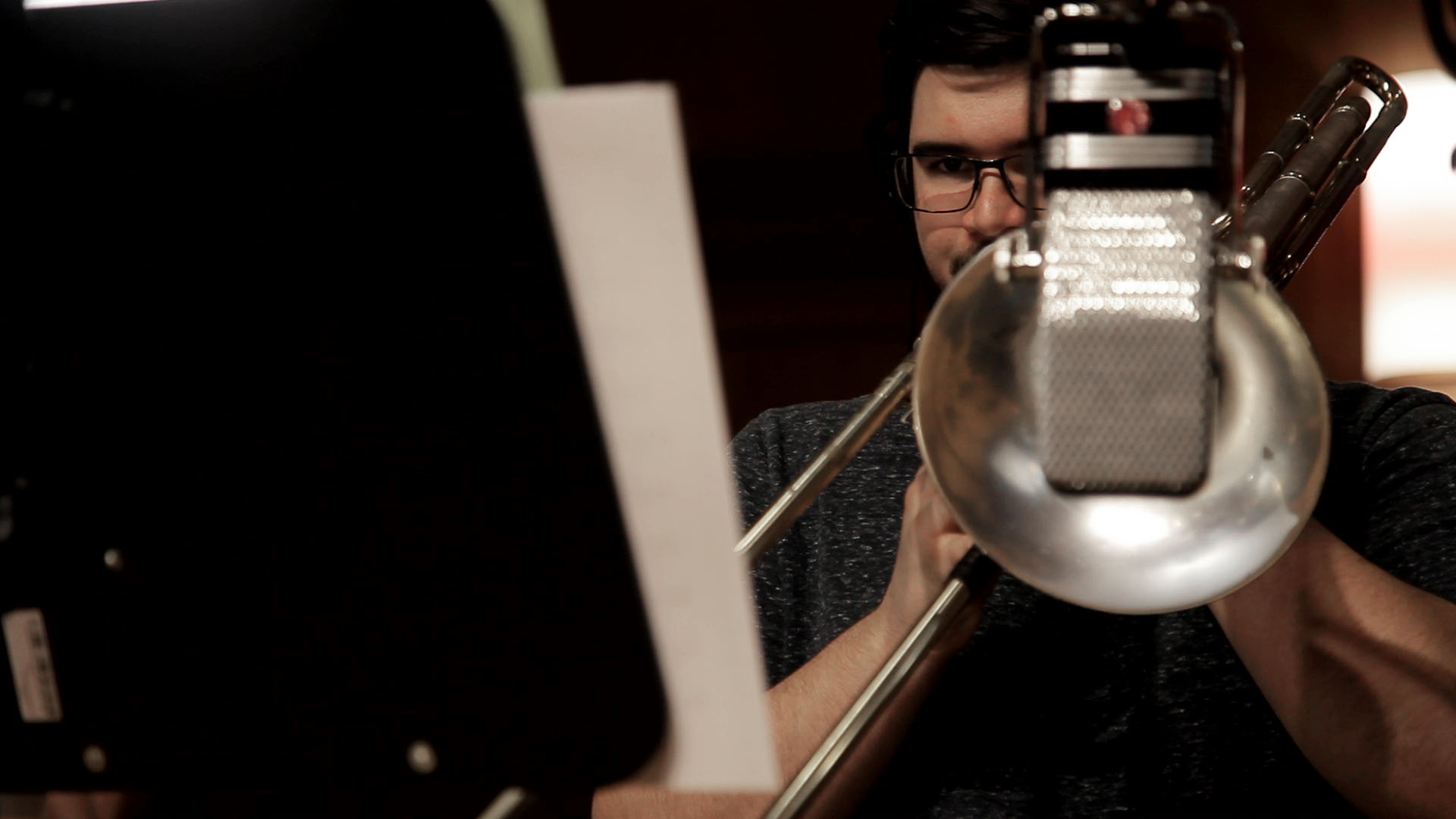 Male musician wearing glasses playing trombone into a microphone with sheet music on a stand in a professional recording studio.