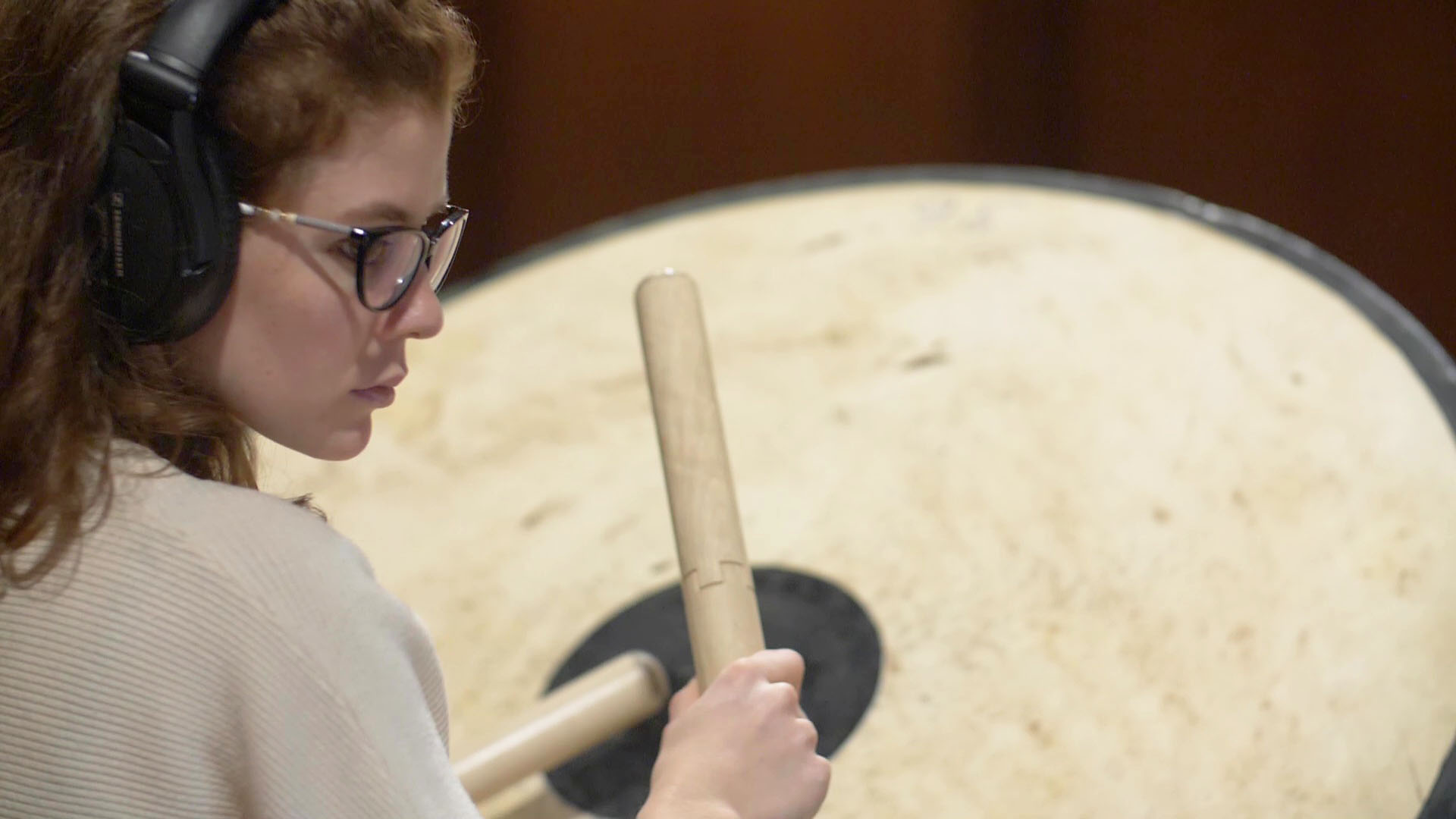 Young woman with headphones and glasses playing a large bass drum with mallets, focused on percussion during a music recording session or performance.
