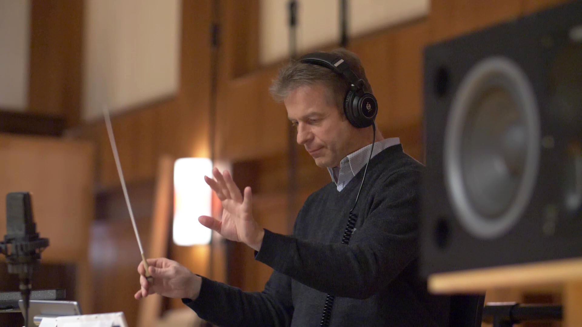 Orchestral conductor wearing headphones, holding a baton, and gesturing with his left hand during a recording session in a studio.