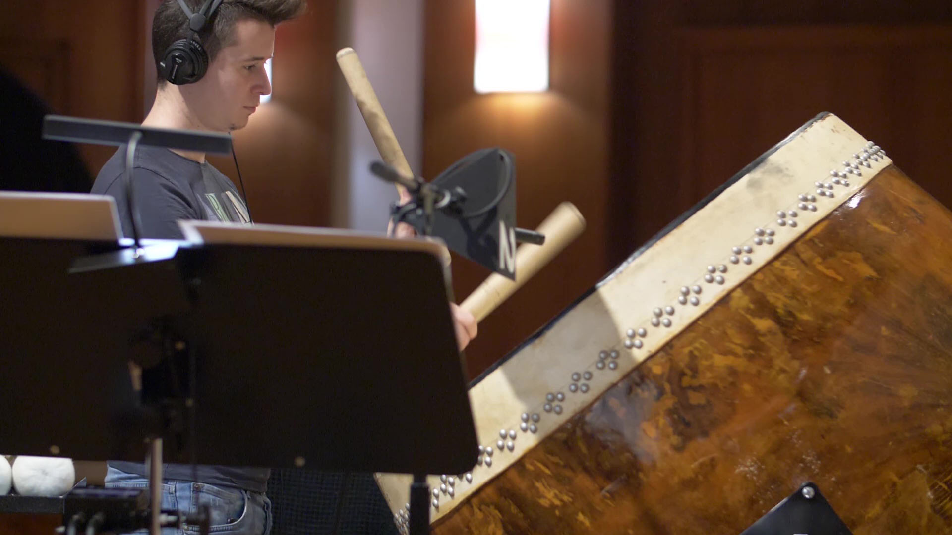 Musician wearing headphones, playing a large taiko drum with drumsticks in a recording studio, featuring a microphone setup.