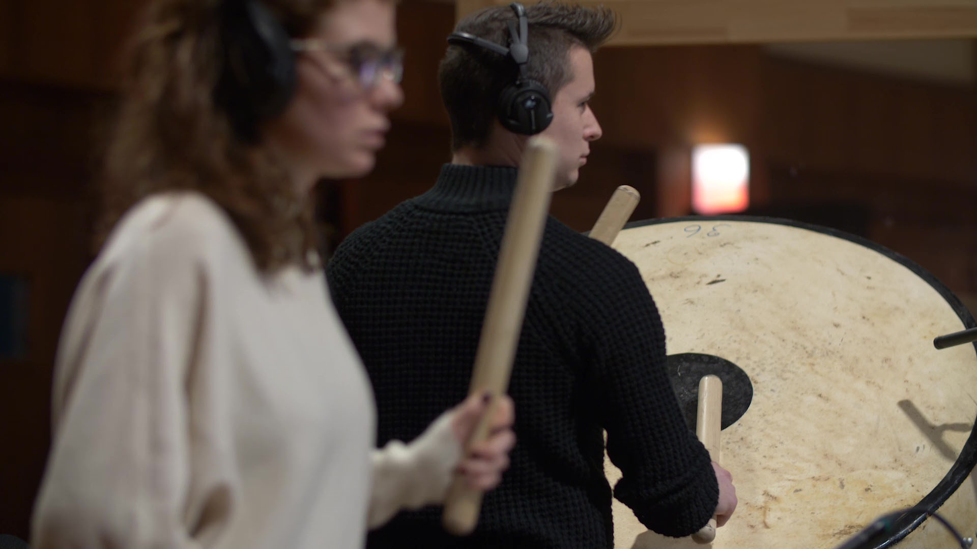 Musicians in a recording studio, a man in black sweater playing timpani drums, a woman with headphones holding drum mallets ready to play.