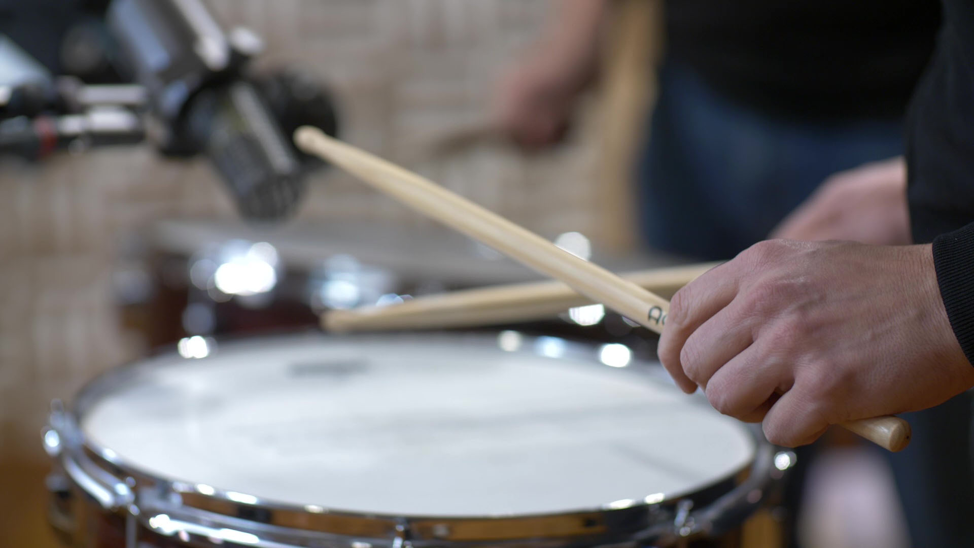 Close-up of a drummer playing a snare drum with drumsticks, a microphone positioned above.