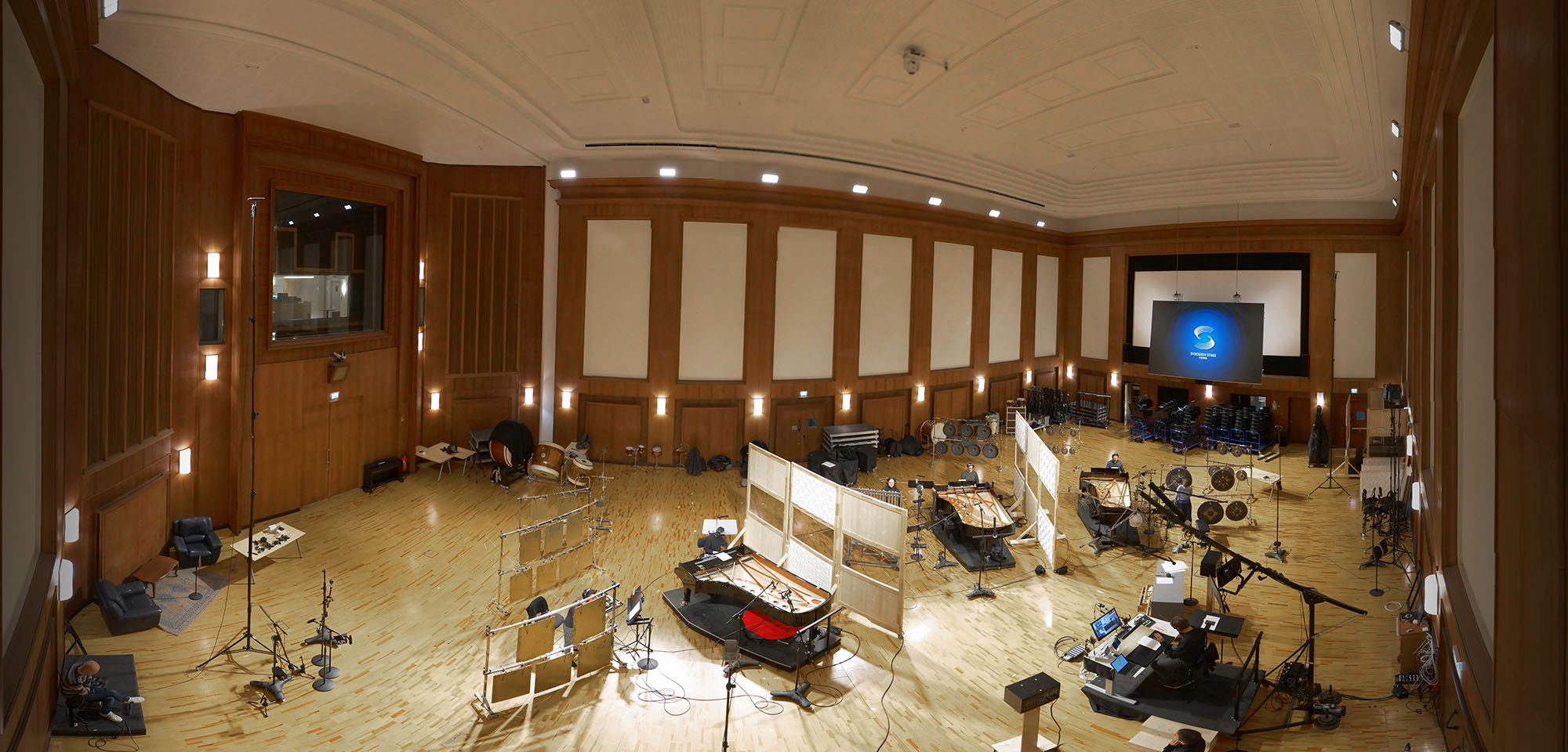 Panoramic view of a large, high-ceiling recording studio with light wood floors. Various musical instruments including grand pianos, percussion, and string instruments are arranged on the floor, surrounded by acoustic panels, microphones on stands, and recording equipment. A large display screen with the Synchron Stage Vienna logo is visible at the far end of the studio.