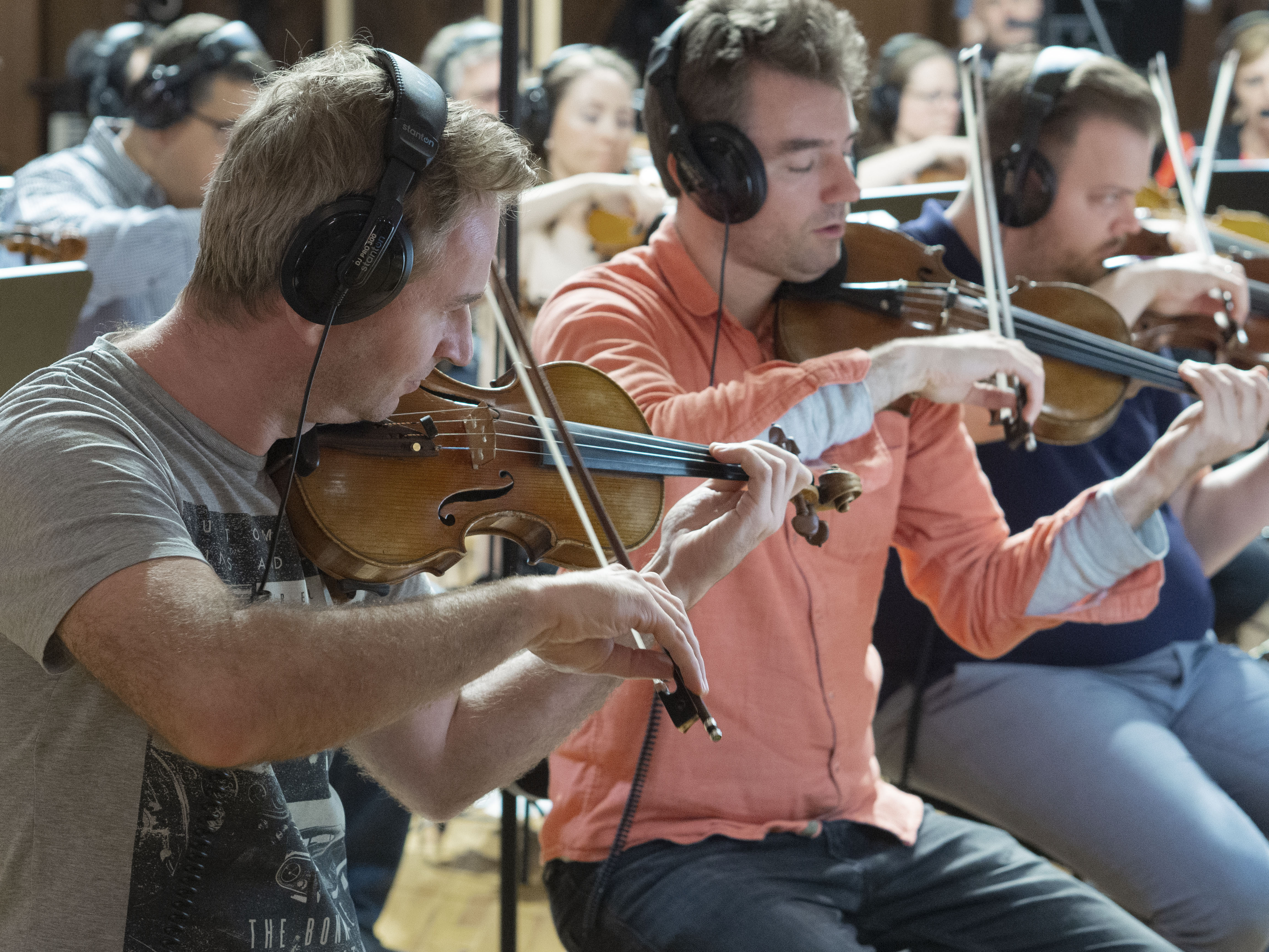 Musicians wearing headphones playing violins in an orchestra during a music production or recording session.
