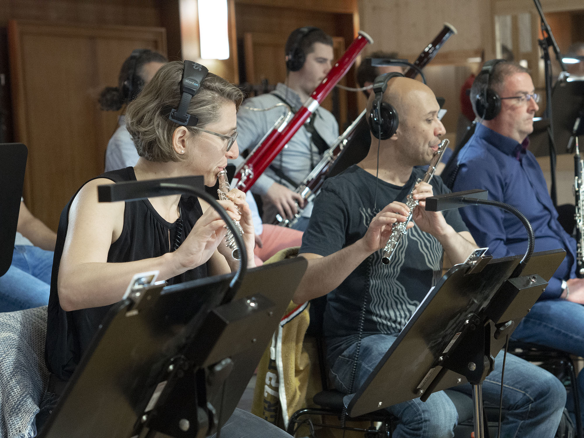 Musicians wearing headphones play flutes and bassoons in an orchestra rehearsal.