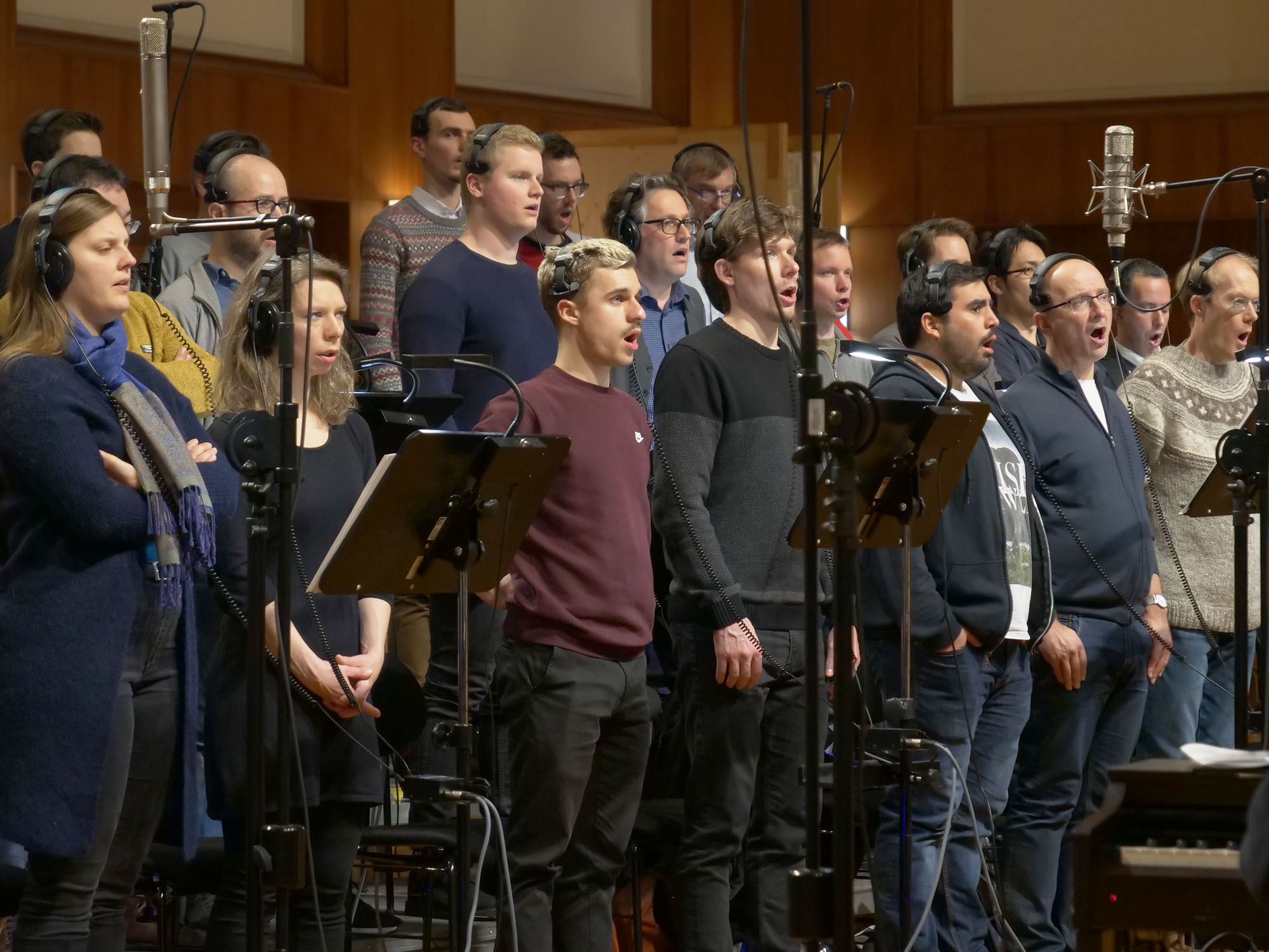 A group of diverse male and female choir members, wearing headphones and standing in front of microphones, sing during a recording session in a studio.