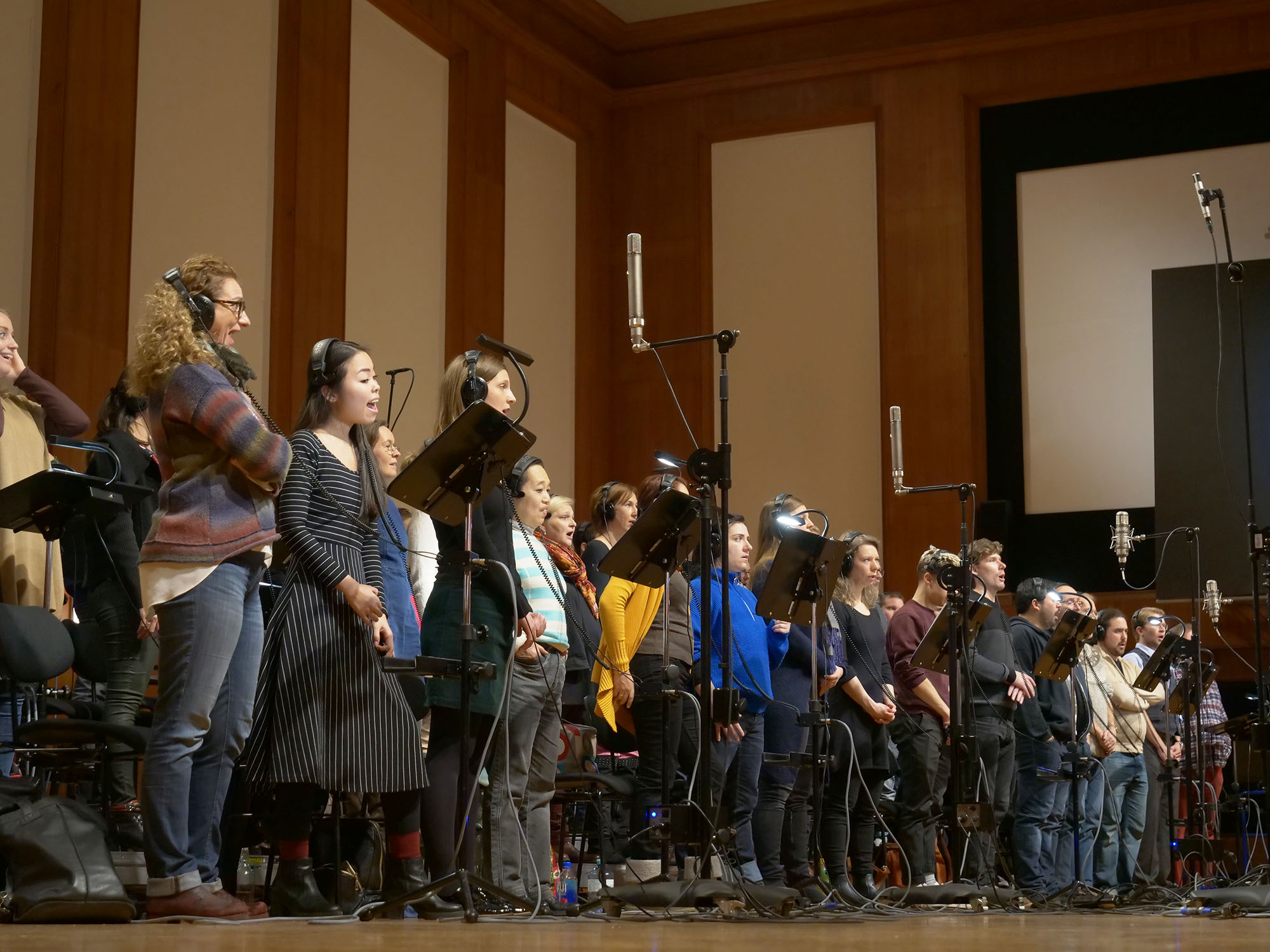 A diverse, large choir singing with headphones on and microphones on stands in a professional recording studio or concert hall.