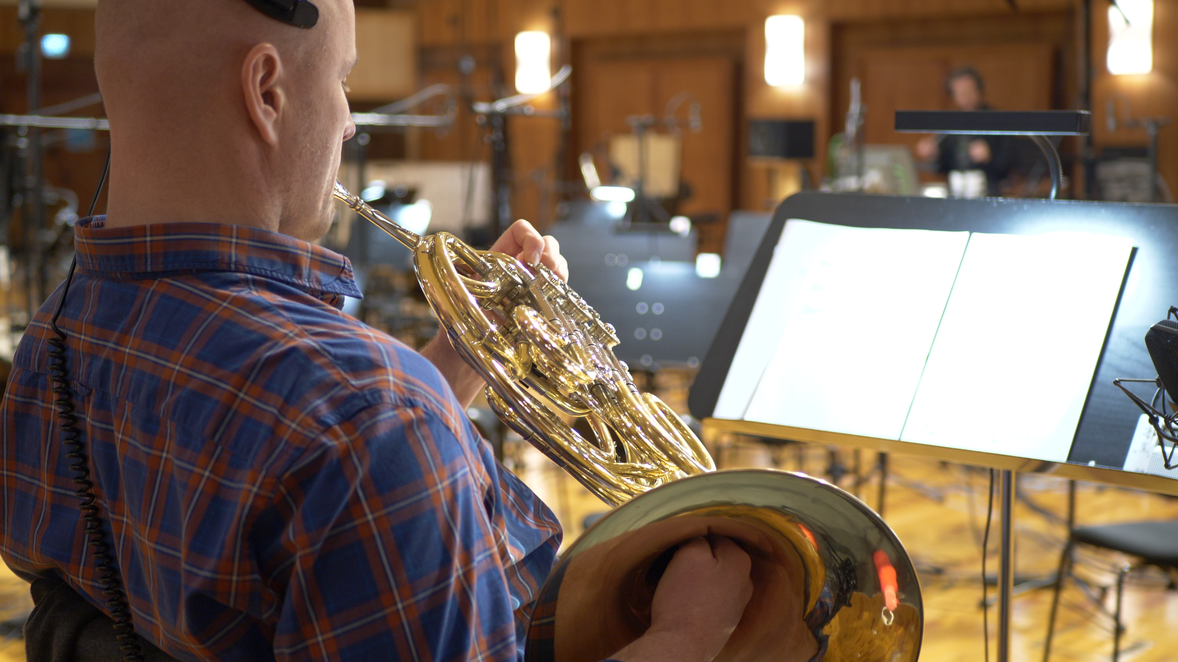 Musician playing a shiny brass French horn with a light brown and blue plaid shirt, music stand and recording studio background.