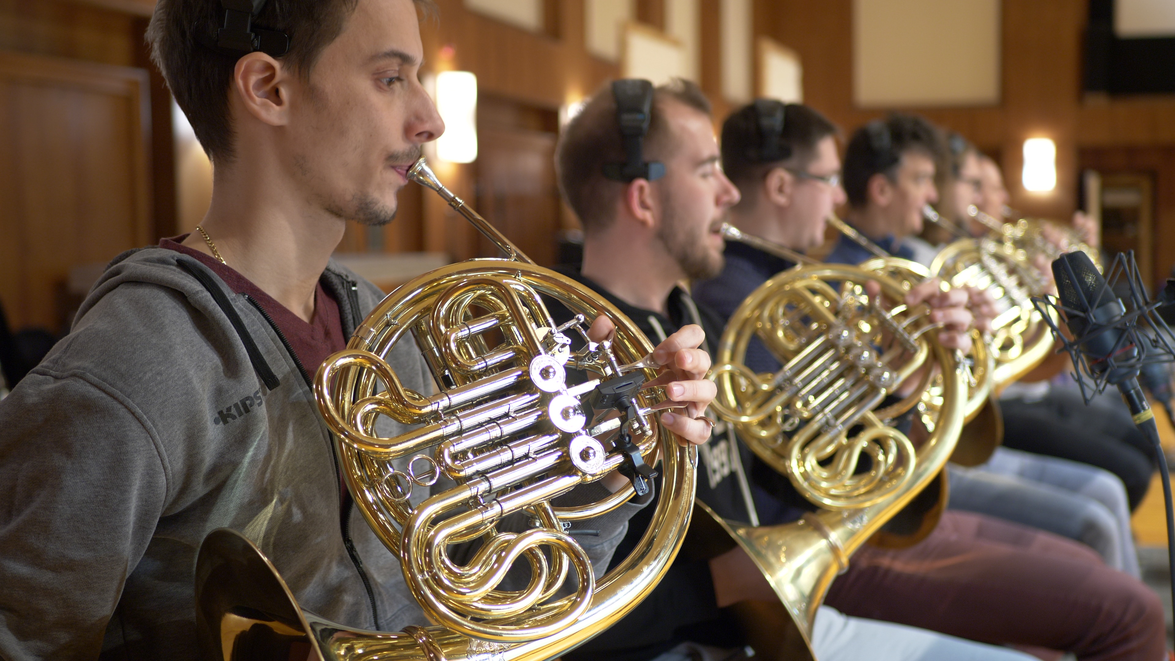 Musicians playing golden French horns in an orchestra practice room, with microphones recording.