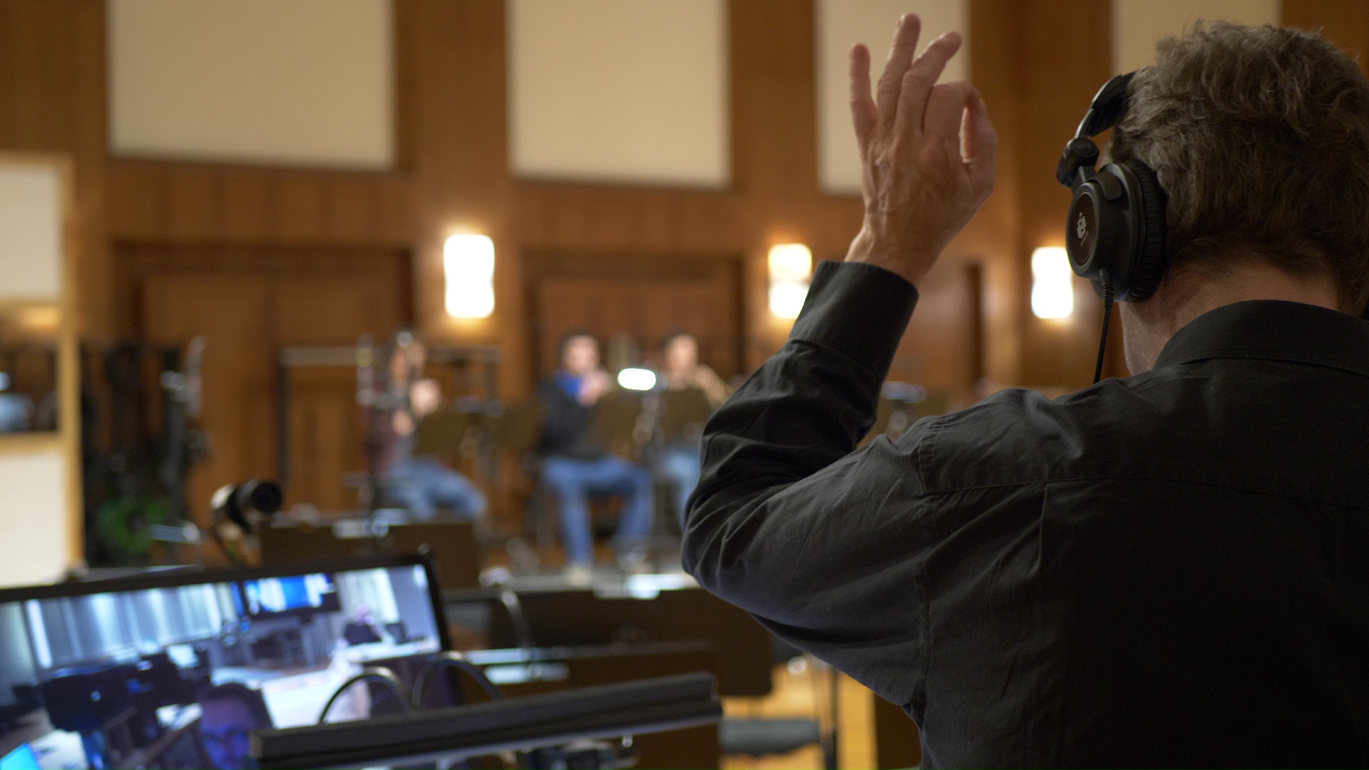 Sound engineer with headphones and dark shirt conducting musicians in a recording studio, monitor showing audio mixing interface, music production.