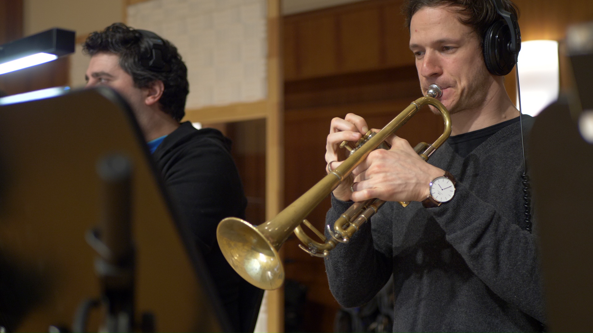 Musician with headphones playing a brass trumpet in a recording studio, another musician blurred in the background.