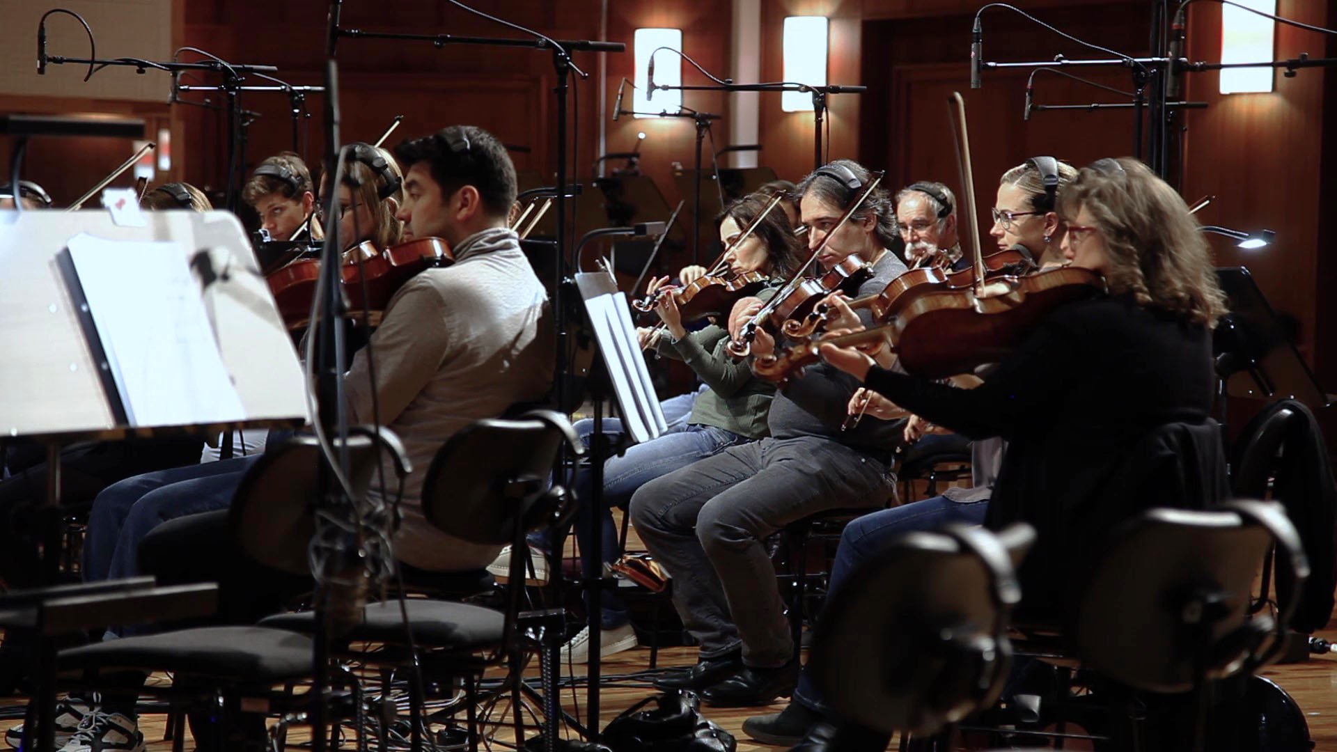 Musicians playing violins in an orchestra with microphones and music stands visible.