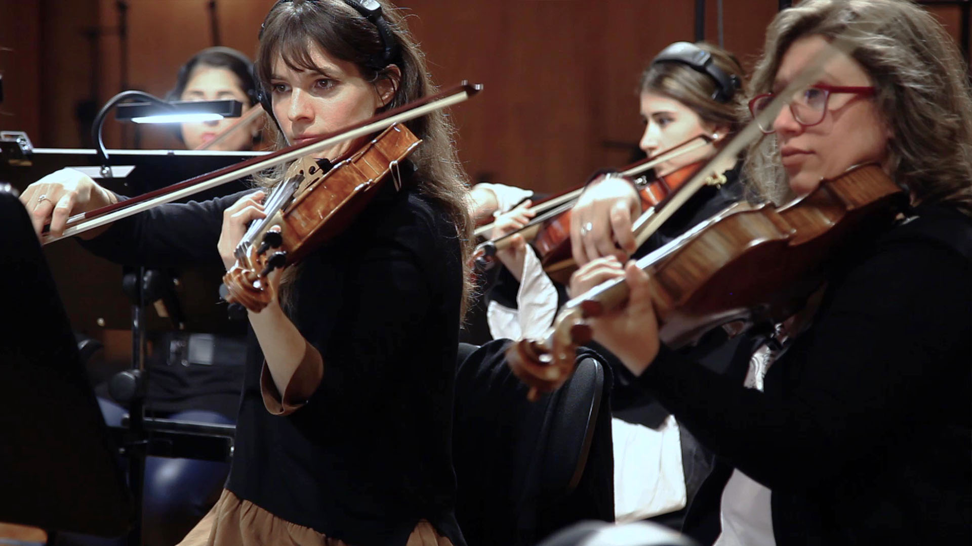Musicians in an orchestra playing violins, with a focus on a woman in the foreground wearing headphones.