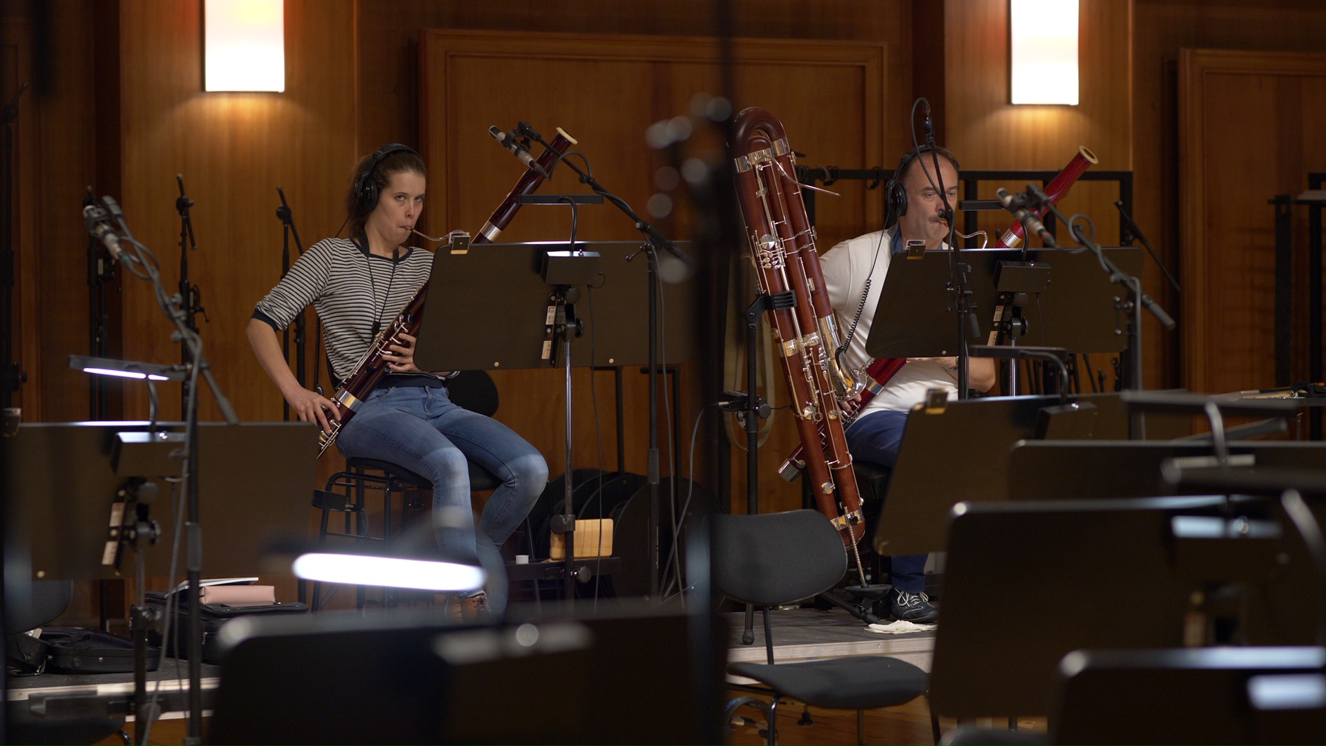 Two musicians, a woman and a man, playing bassoons during a recording session in a studio or concert hall.