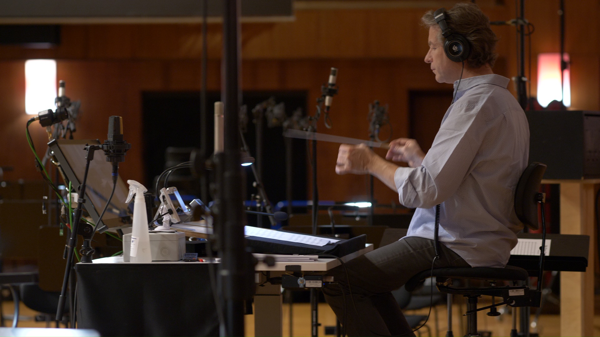 Composer or conductor wearing headphones, seated at a desk with music scores, surrounded by microphones and recording equipment in a professional sound studio, actively working on an audio production.