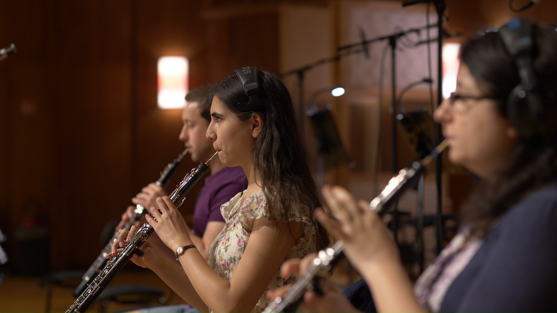 Three musicians, two women and one man, playing oboes in a recording studio. The central woman wears headphones and a floral top, actively performing. Microphones and music stands are visible in the background, suggesting a professional recording session.