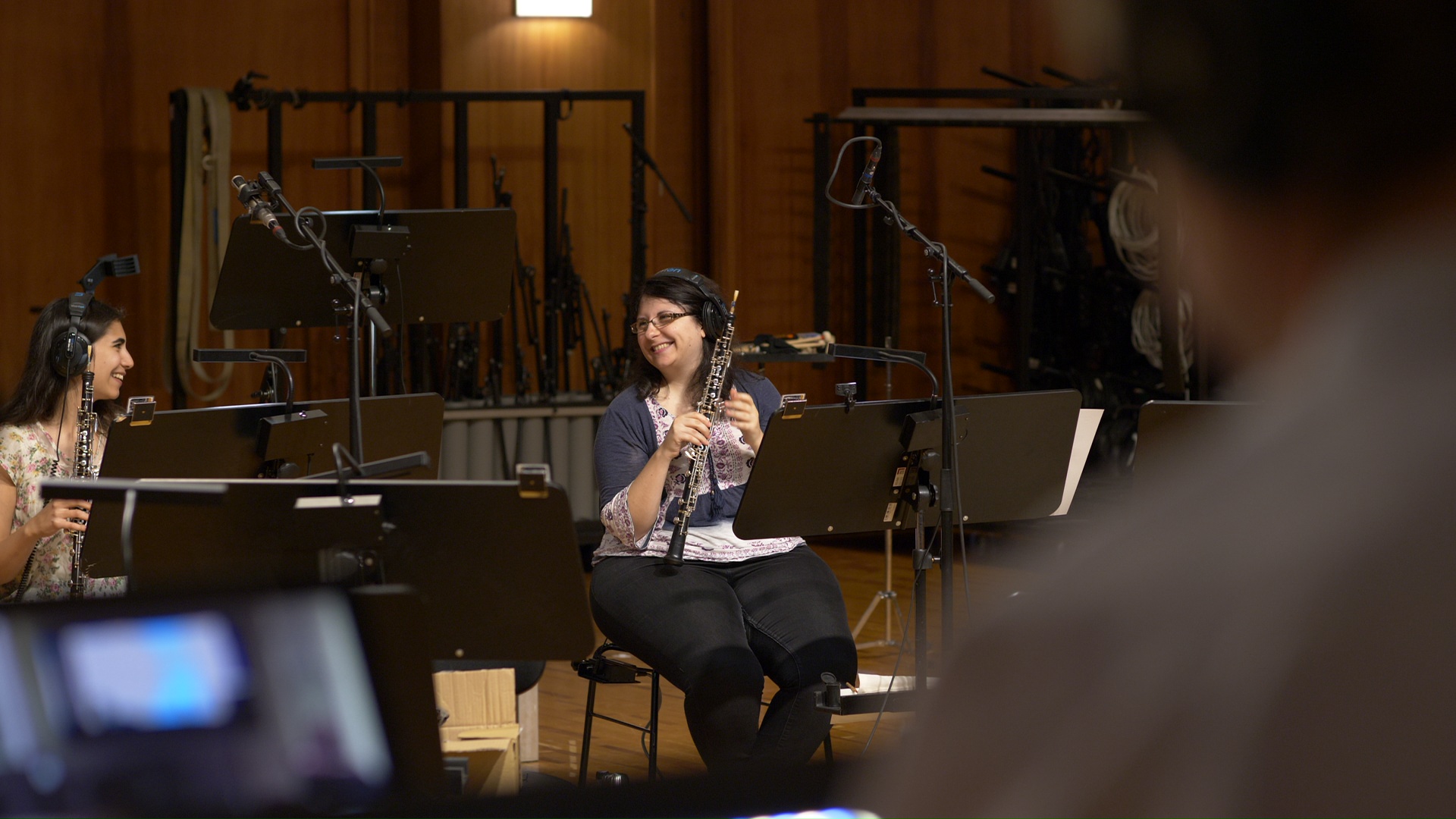 Two female musicians, one young and one older, happily playing oboes while wearing headphones in a recording studio with microphones and music stands.