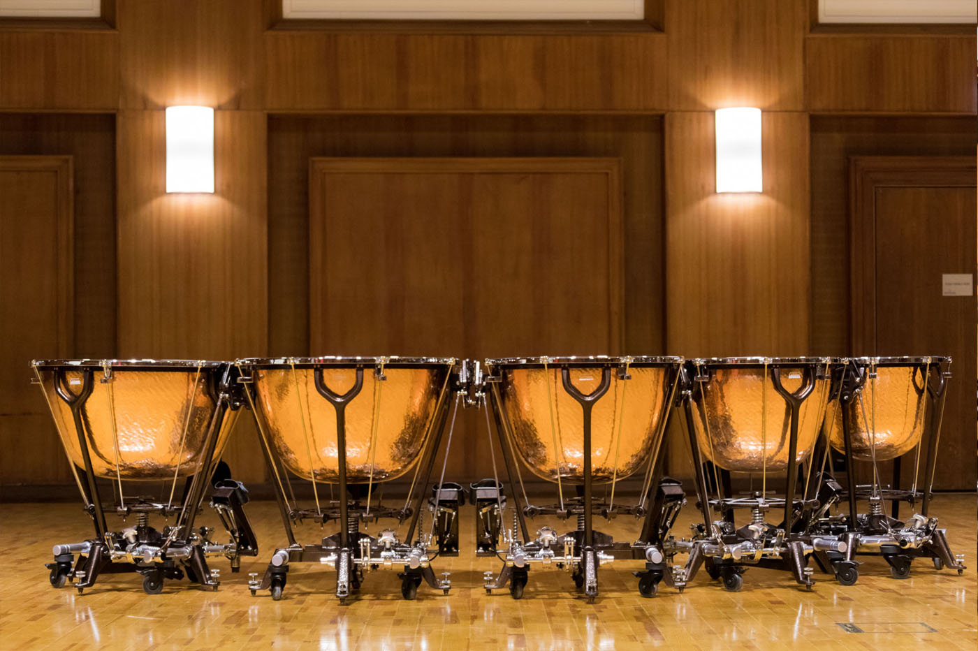 A row of five polished copper timpani drums with detailed stands on a wooden stage, with wood-paneled walls and two lit sconces in the background.