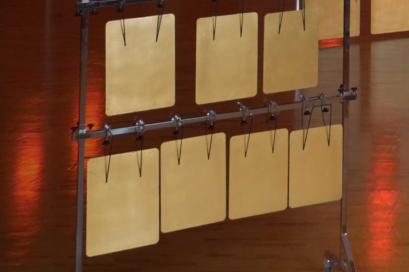 Two rows of golden square plates, part of a percussion instrument, hang from a metal frame on a stage with warm lighting.