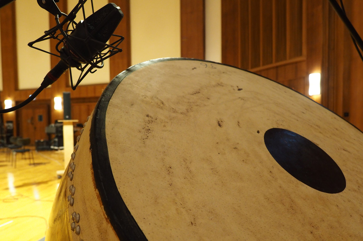 Studio microphone positioned above a large drum or timpani in a recording room with wooden walls and flooring, ready for musical performance or sound recording.