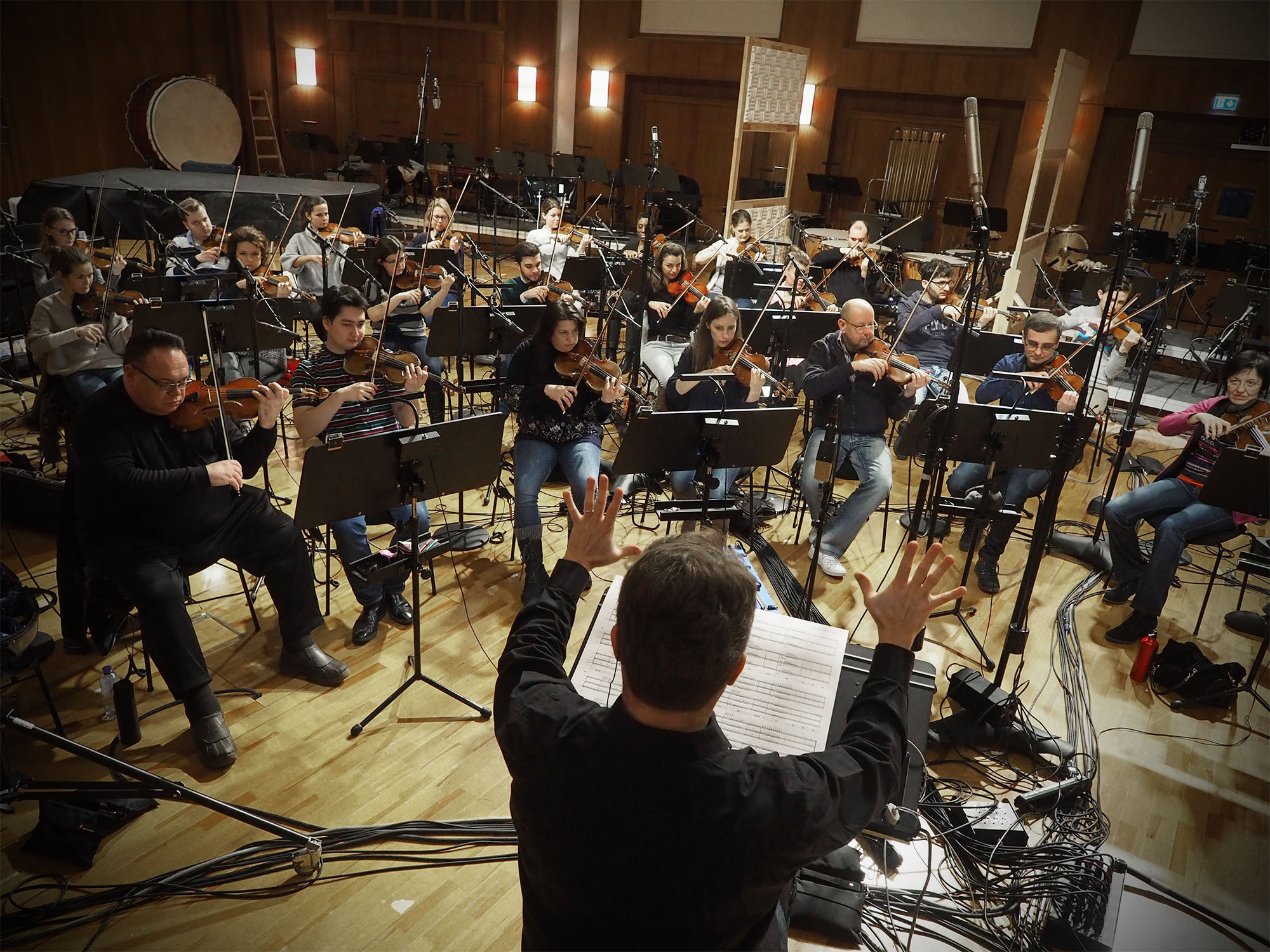 Orchestra conductor directing musicians playing violins in a recording studio, featuring professional audio equipment and a large bass drum.