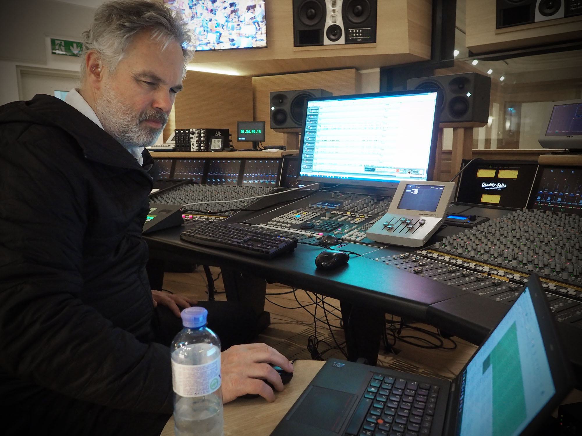 An audio engineer with grey hair and beard, wearing a black jacket, sits at a large mixing console with multiple screens, speakers, and sound equipment. He is looking down intently at a black laptop positioned next to a water bottle on the desk, while operating a computer mouse with his right hand. The background shows professional studio monitors and various other recording studio components.