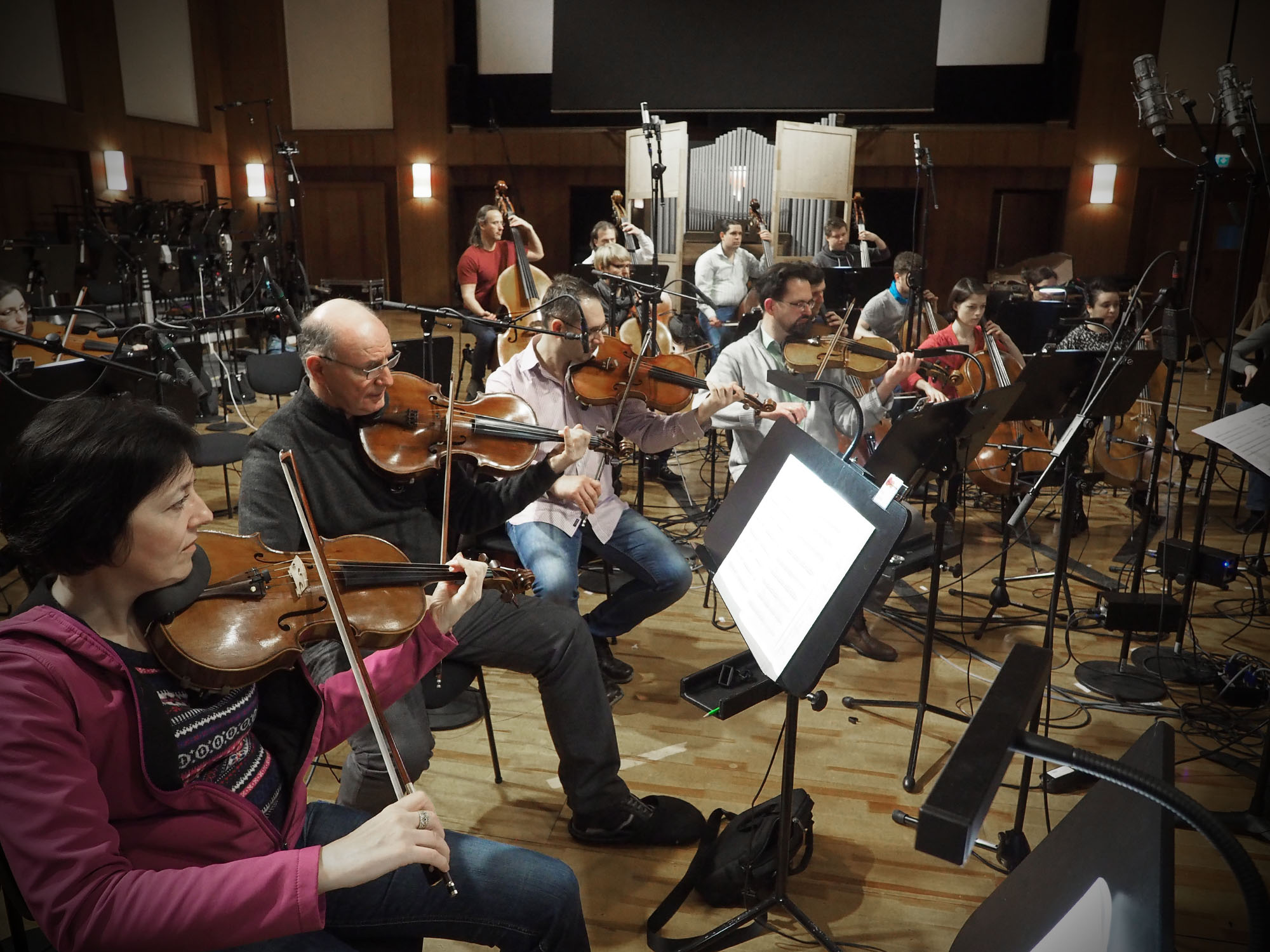 Orchestra musicians performing in a recording studio with string instruments, music stands, and microphones.