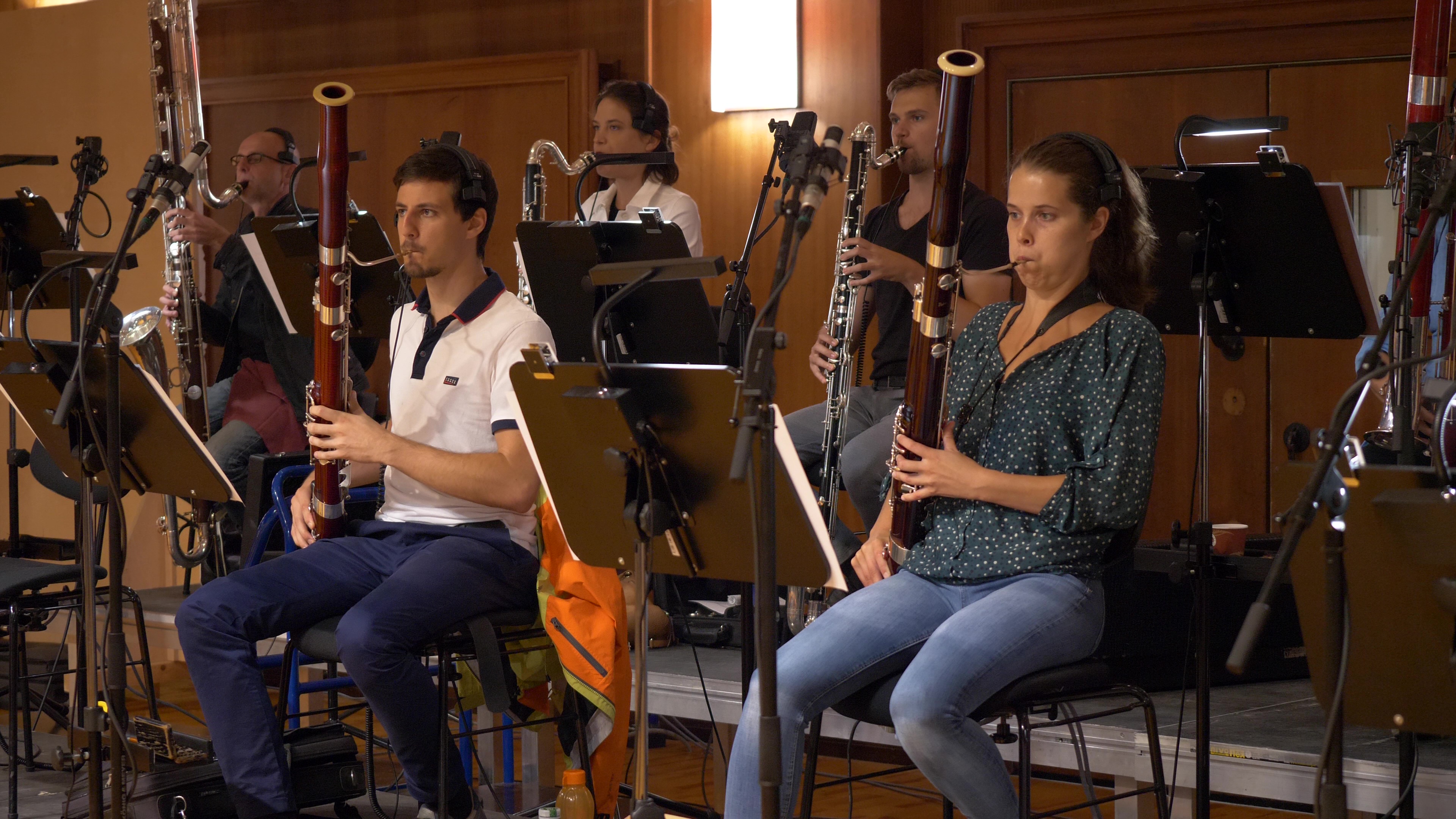 Ensemble of five musicians playing various bassoons and contrabassoons, seated with sheet music and microphones in a recording studio.