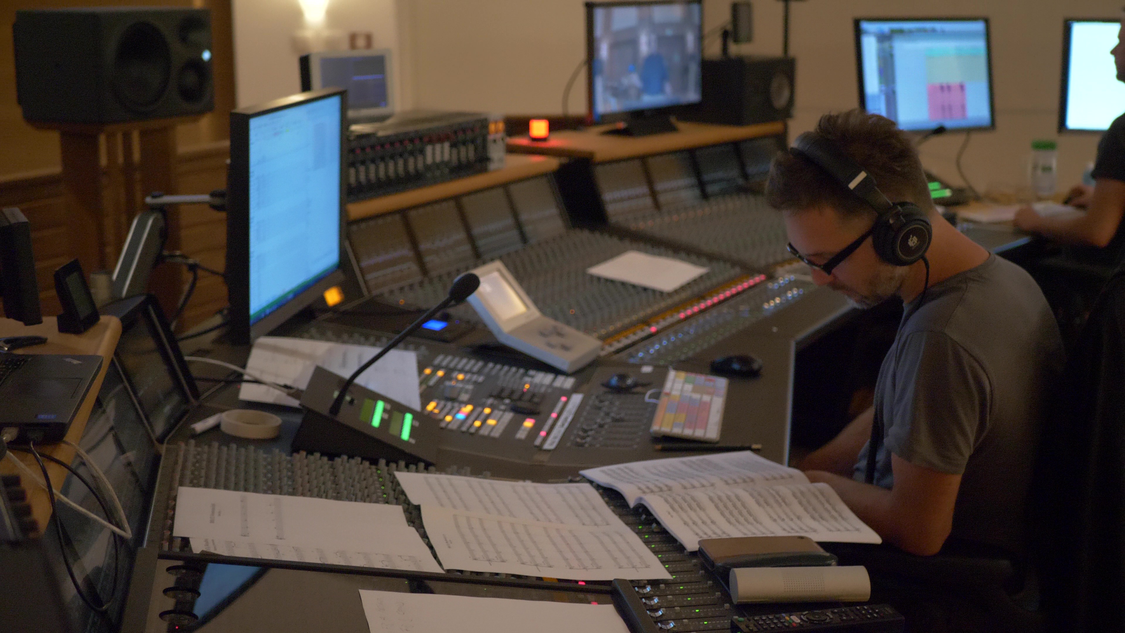 Audio engineer in a modern sound studio wearing headphones and glasses, looking at sheet music over a large mixing console with multiple screens displaying audio software.