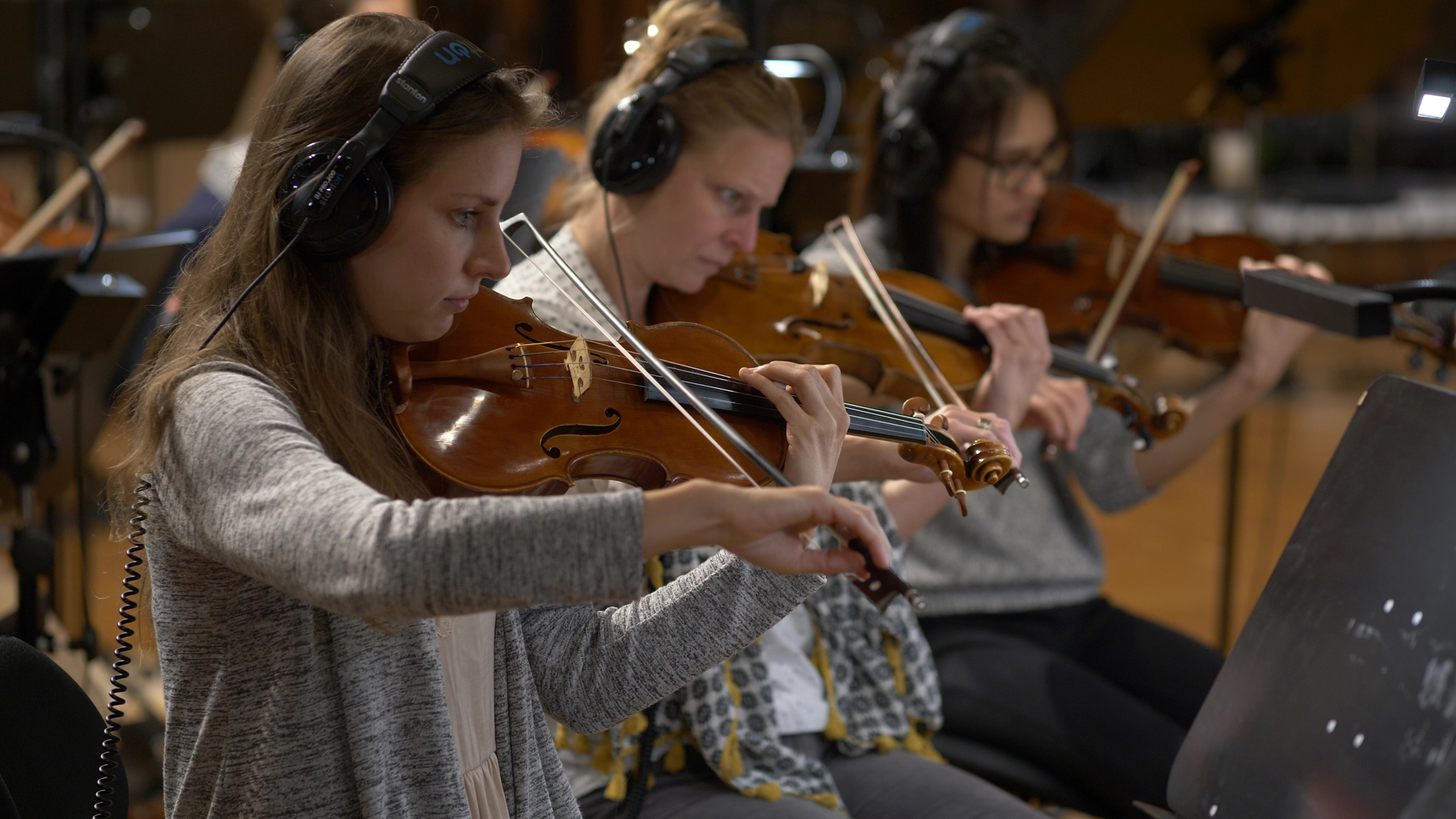 Three female musicians wearing headphones playing violins in an orchestra or recording studio.